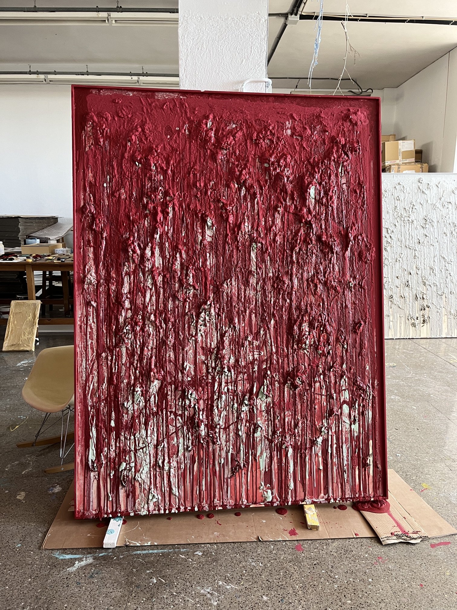 Cuadro de arte abstracto con pintura en relieve en tonos rojos y blancos, en un espacio de taller o estudio.
