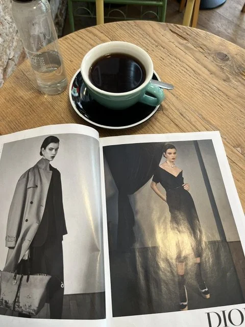 Una taza de café negro sobre una mesa de madera, acompañada de un libro abierto con fotografías de moda y una botella de agua.