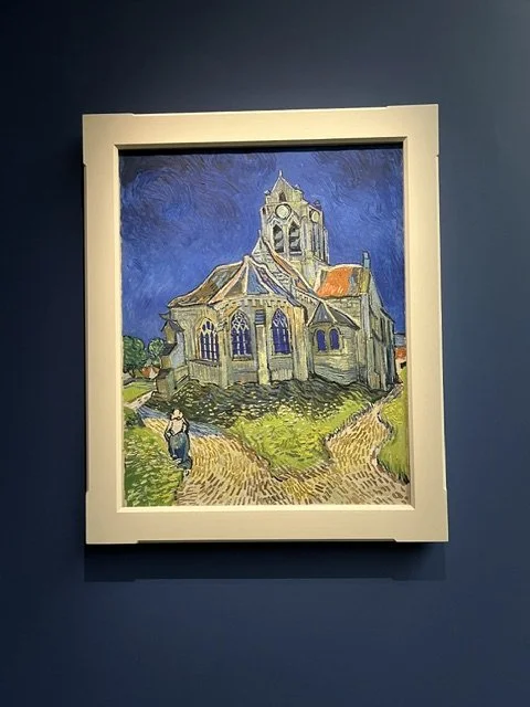Una pintura de una iglesia antigua con un cielo azul en un marco blanco, en una pared azul.