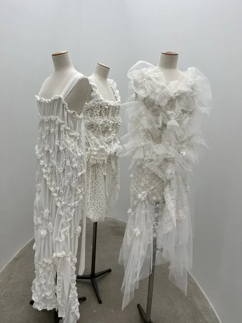 Tres maniquíes con vestidos blancos de estilo floral y con volantes, en exhibición en una pared blanca.