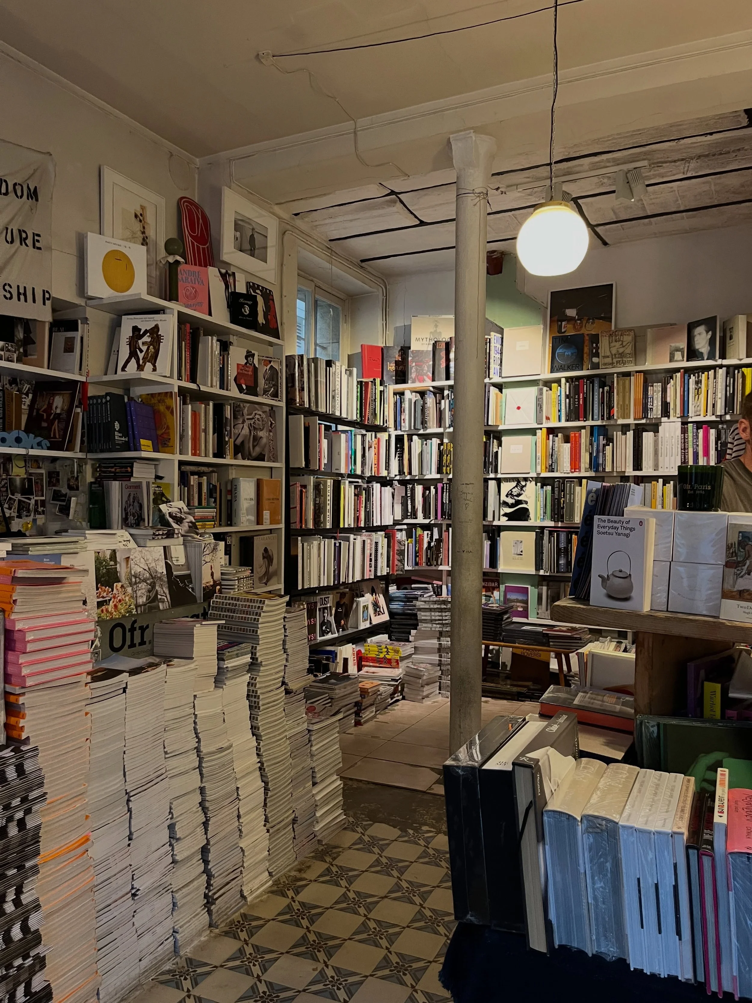 Estanterías llenas de libros en una librería o tienda de libros, con pilas de libros en la mesa y en el suelo, en un ambiente de interior con iluminación cálida.