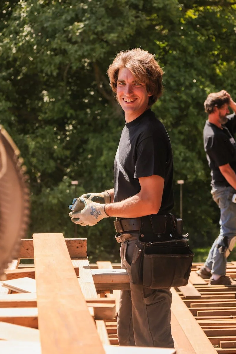 Young man smiling at construction site, holding tools, with another worker in the background, surrounded by wooden beams and trees.