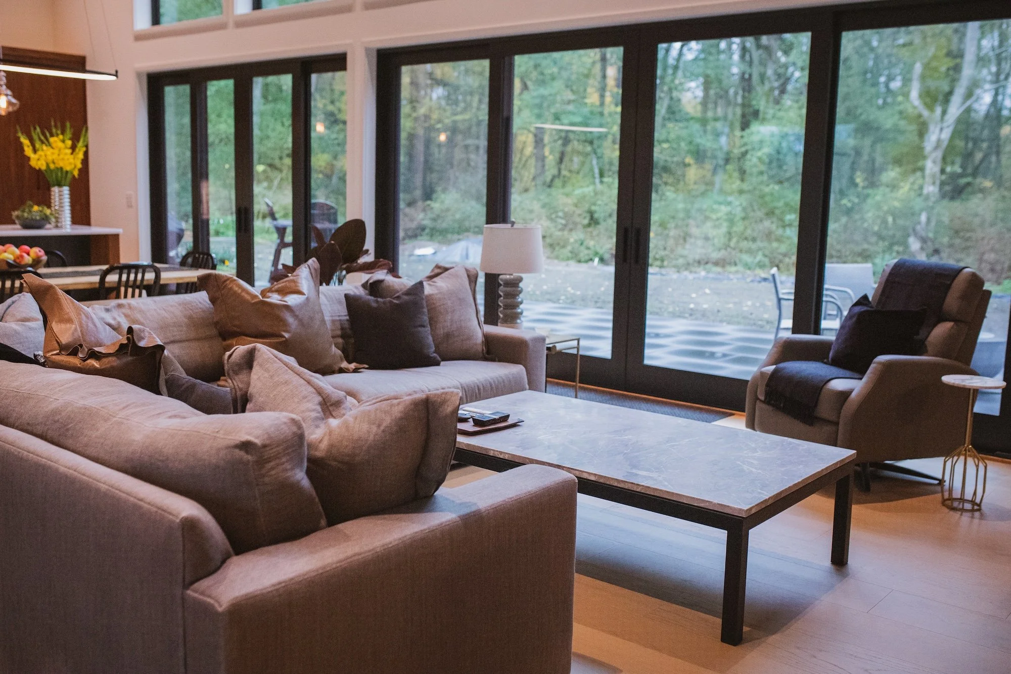 A living room with a beige sectional sofa, a gray armchair, a white table lamp, and large glass sliding doors showing a wooded outdoor area.