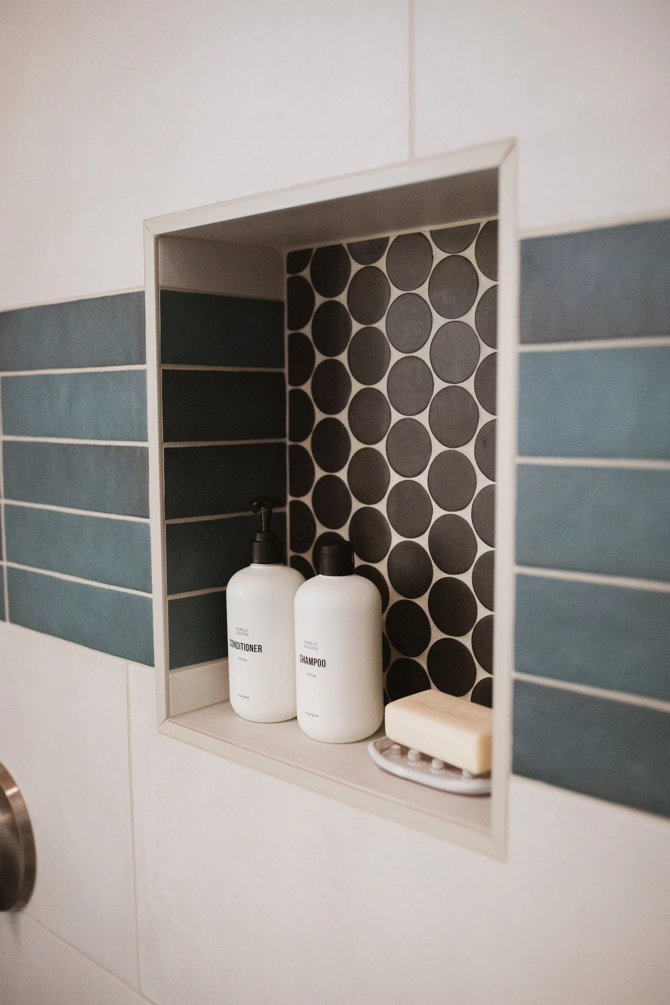 Close-up of a built-in bathroom shelf with two white bottles labeled 'Conditioner' and 'Shampoo', a soap dish with soap, and a decorative black and white polka dot background inside a tiled shower alcove.