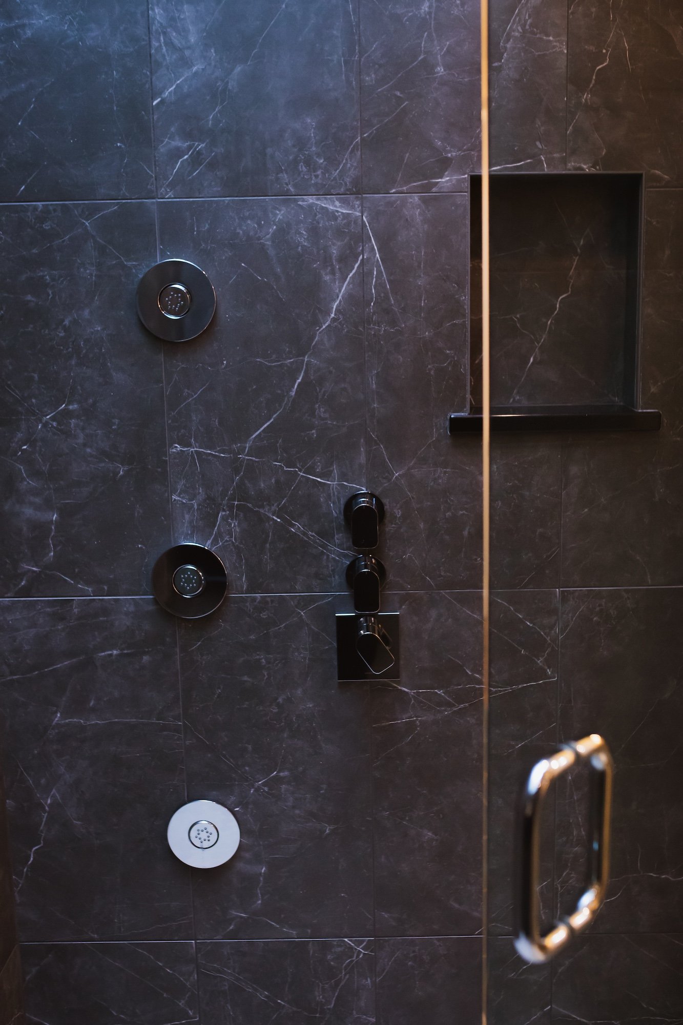 A walk-in shower with dark marble tiles, black shower controls, and a built-in black shelf. The photo is taken from outside the shower door, which has a metal handle visible on the right side.