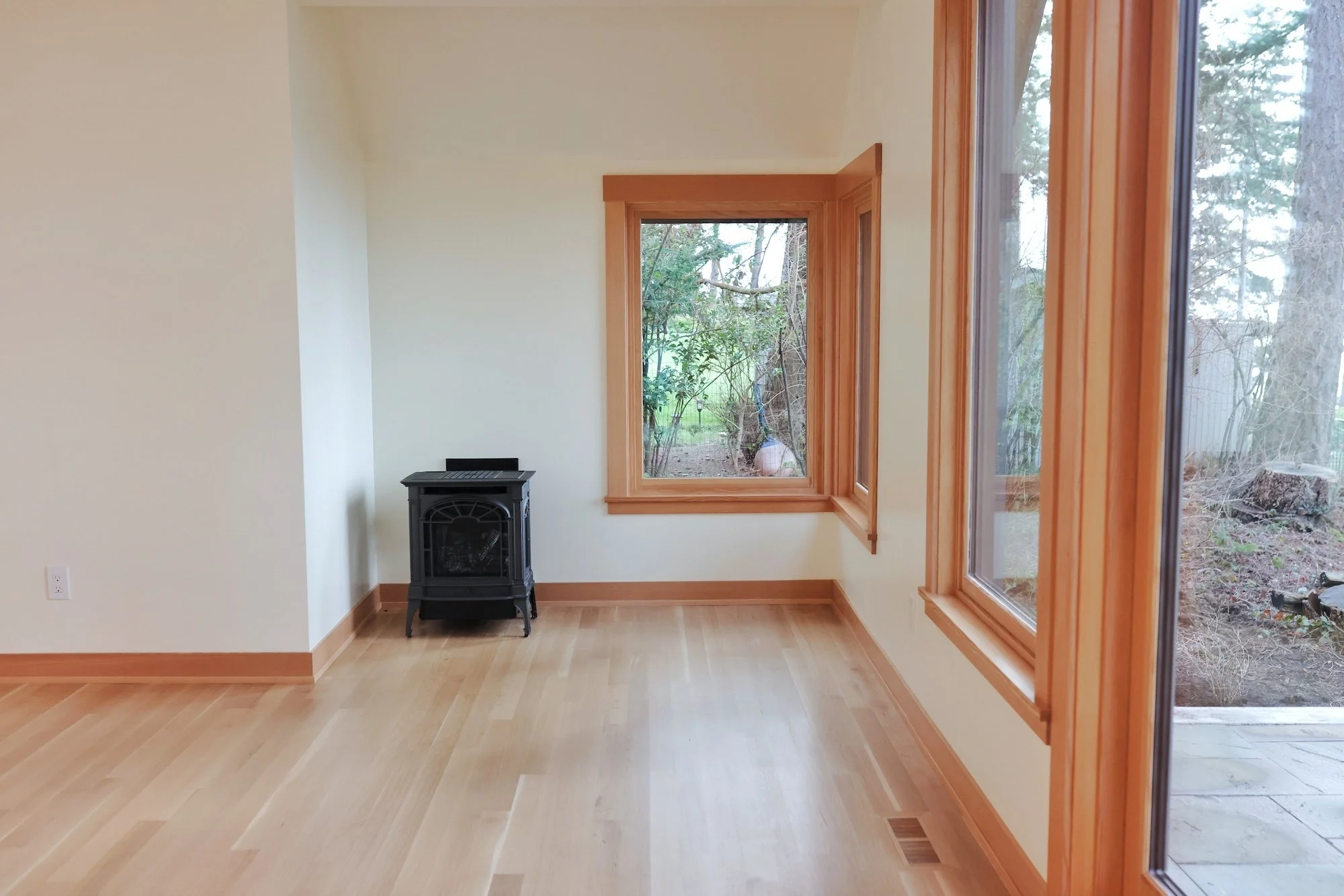 A bright corner of a room with light hardwood floors, cream-colored walls, and large wooden-framed windows revealing outdoor trees and a garden. There is a small black stove or heater near the wall.
