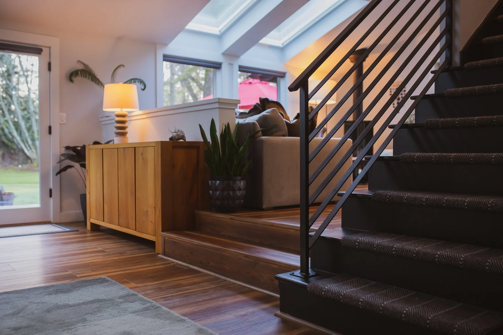 Interior of a modern living room with large skylights, a staircase with black railing, a beige sofa with pillows, a wooden sideboard with a lamp and plants, large windows with blinds, and hardwood floors.