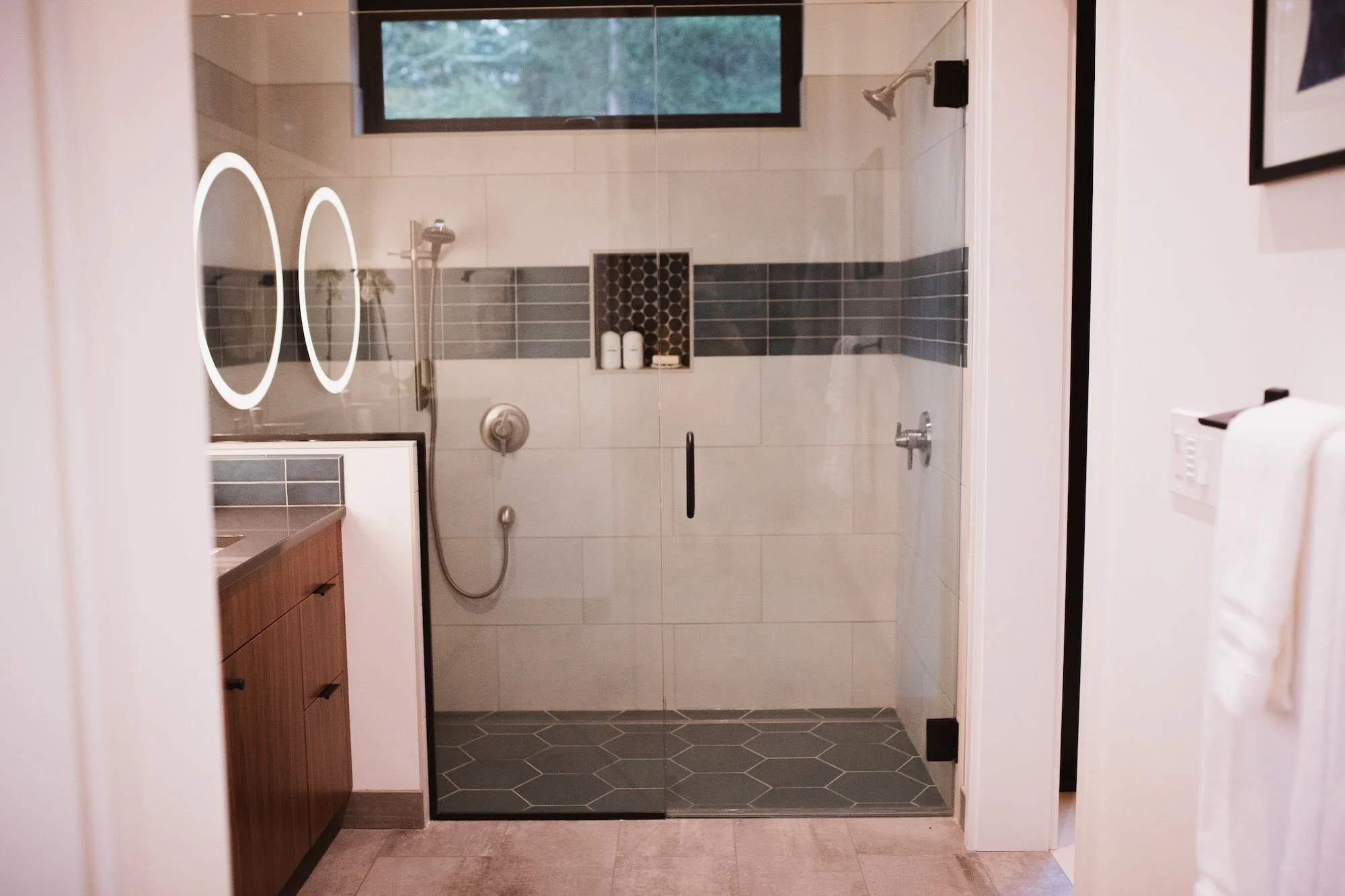 Bathroom with walk-in shower, dark gray hexagonal tiles on the floor, gray and white tiled walls, built-in shelf with bottles, a window with a view of trees, and a wooden vanity with drawers.