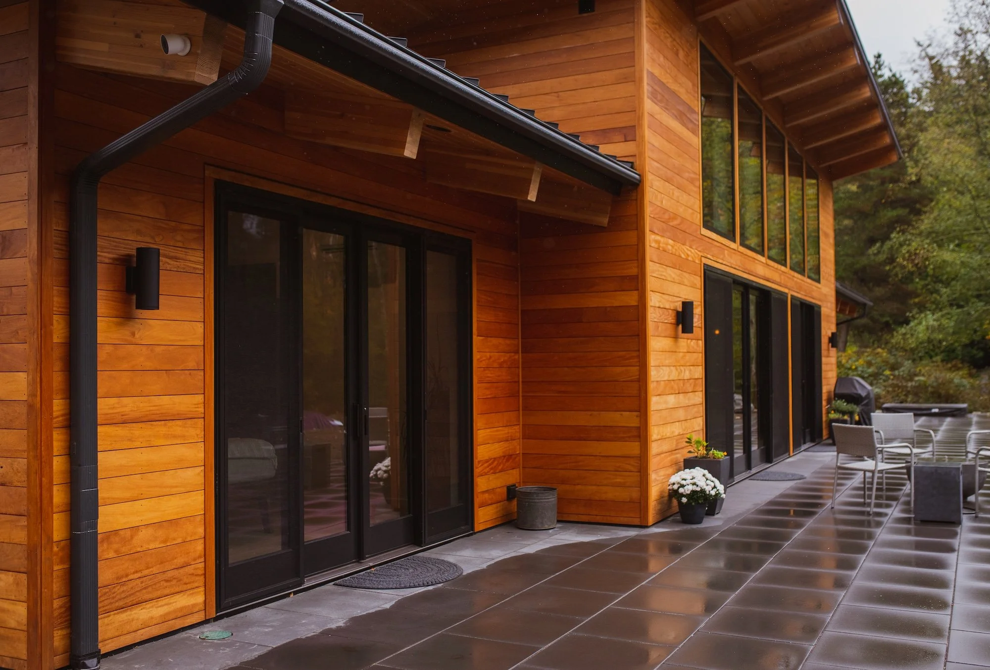 A modern house with wooden exterior walls, large sliding glass doors, a covered patio with outdoor furniture, and a tiled floor. There are trees in the background and rain droplets on the image.