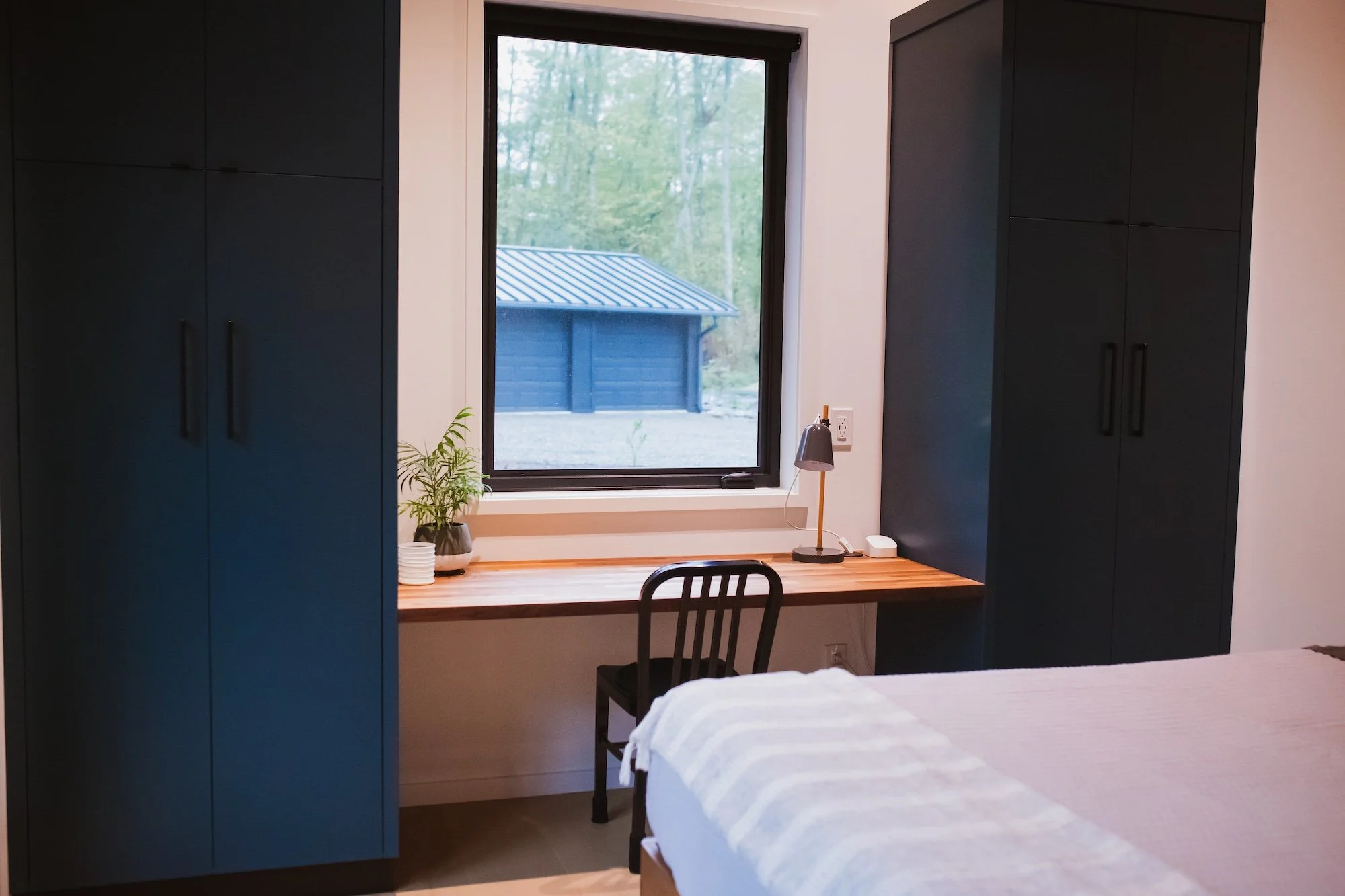 Bedroom corner with a desk below a window showing trees outside, flanked by dark wardrobe cabinets, with a potted plant, a small lamp, and a chair in the room.