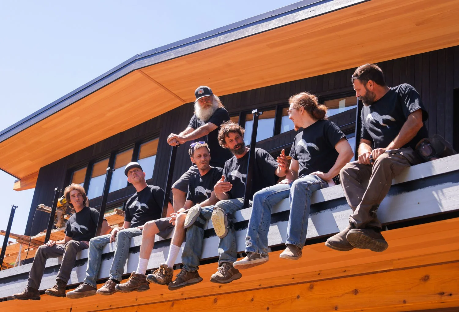 Group of seven people sitting on the edge of a balcony of a modern building, engaged in conversation, with wood and black panels on the exterior and large windows.