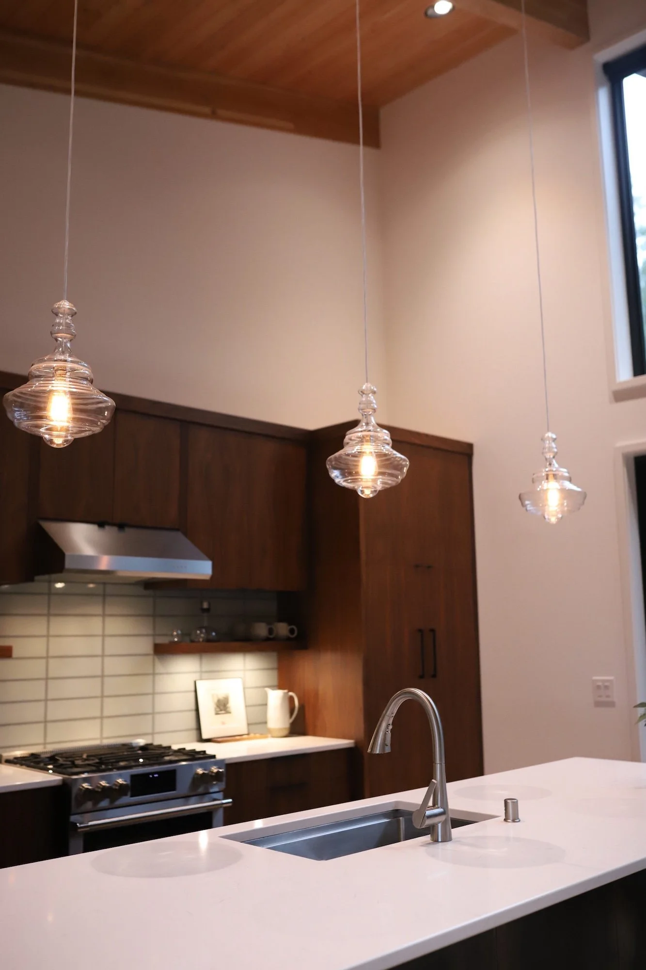 A modern kitchen with a white island countertop, a stainless steel sink with a faucet, and three glass pendant lights hanging from the wooden ceiling. There are wooden cabinets, a stove, and a tiled backsplash in the background.