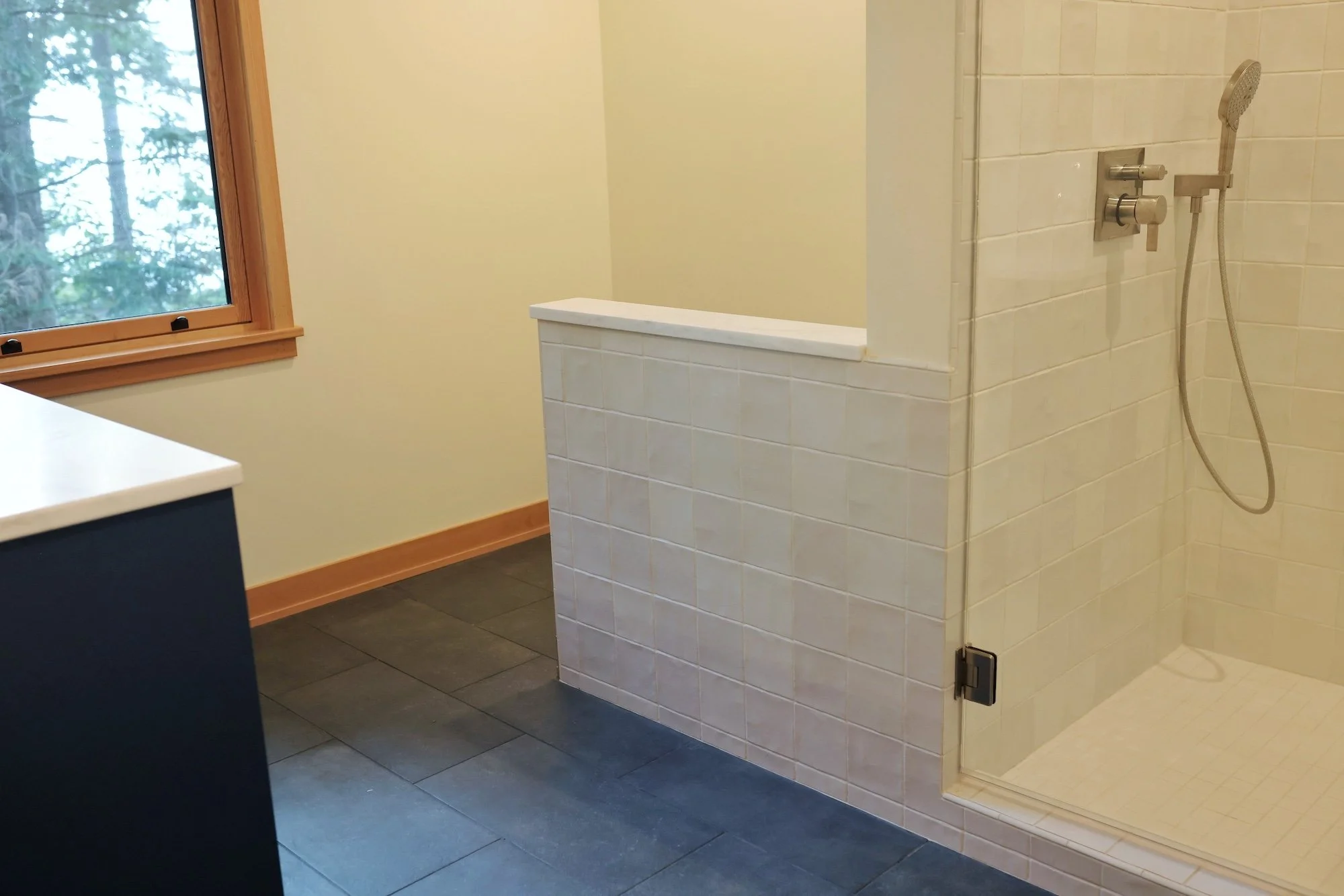 A bathroom with a shower area enclosed by white tiles, a handheld showerhead, a window with a wooden frame, and a black cabinet with a white countertop.