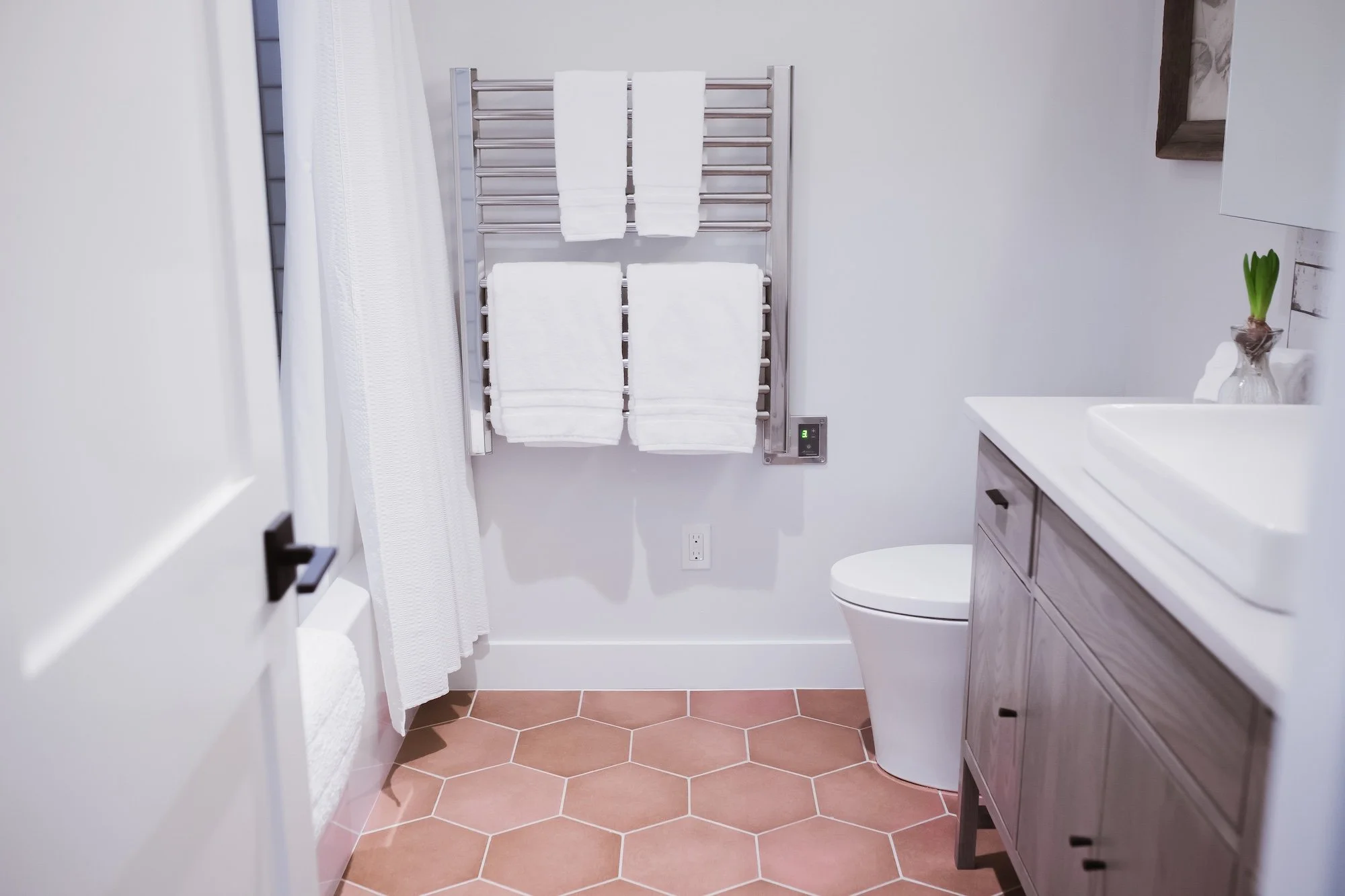 A modern bathroom with a heated towel rail, white towels, a white sink, a brown cabinet, a small potted plant, and a partial white countertop. The floor has hexagonal terracotta tiles.