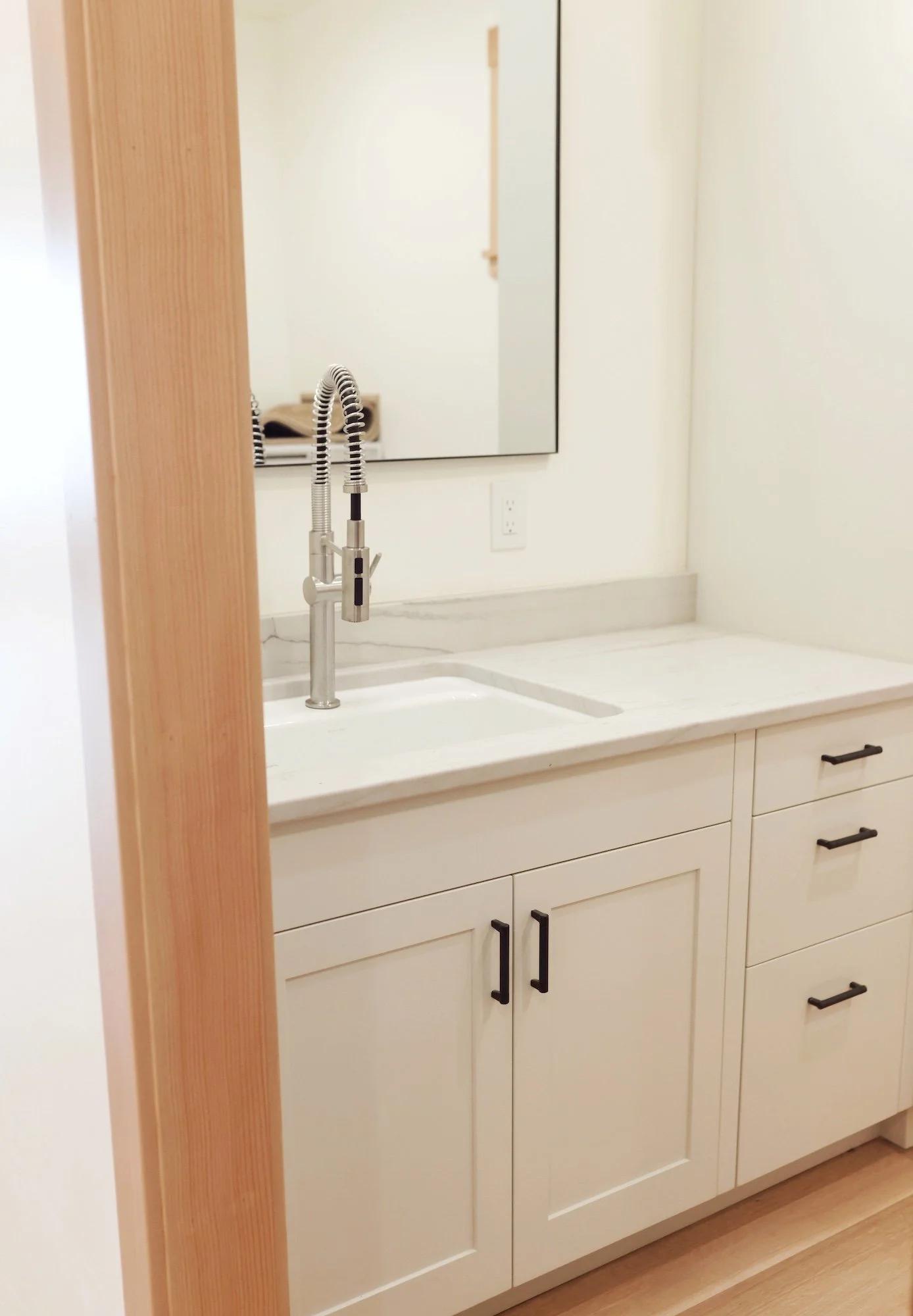 Modern kitchen sink with black handles, white cabinet drawers, a large mirror, and a double-handle faucet with a flexible sprayer, next to a wooden wall.