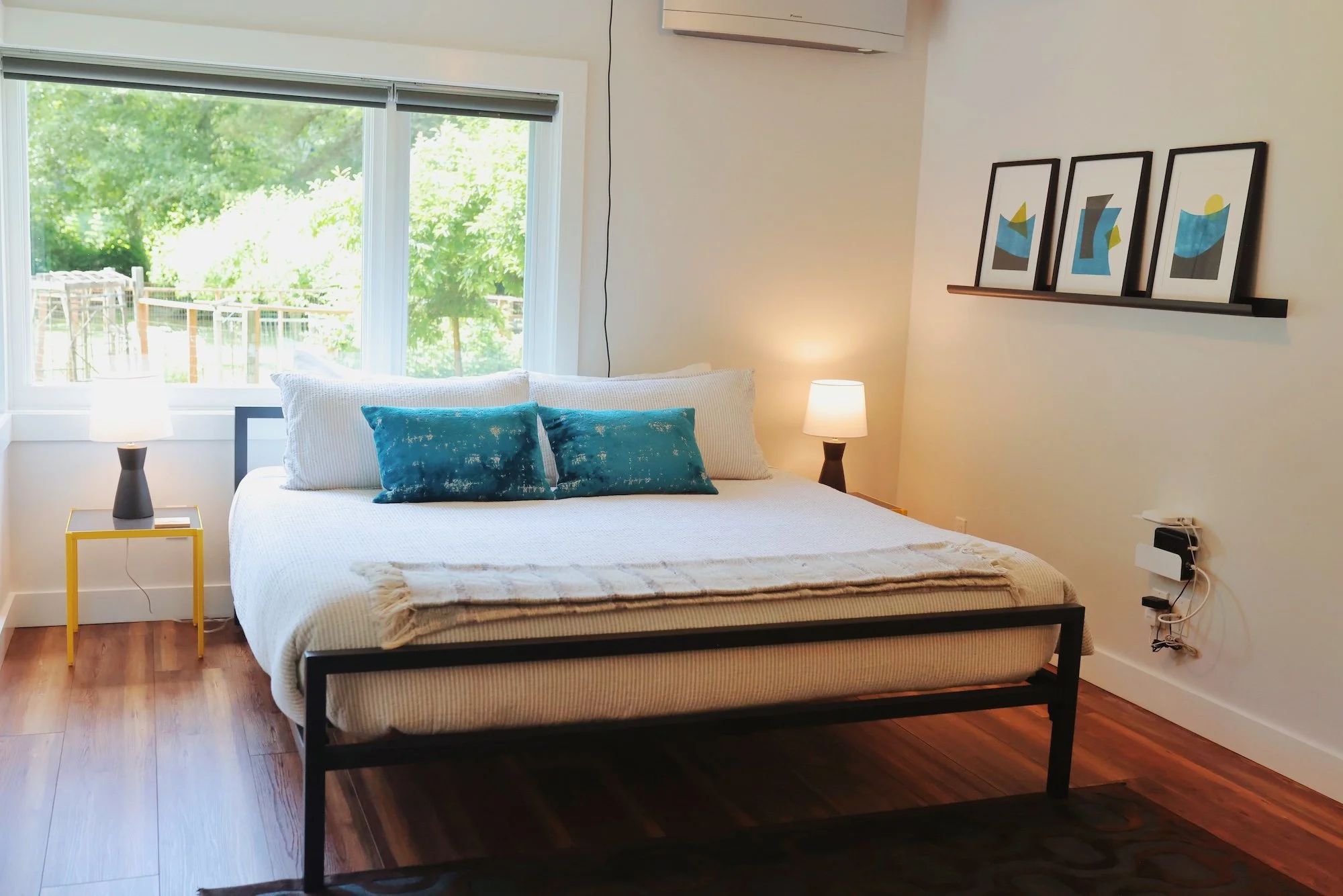 Bedroom with a bed, two teal pillows, white pillows, and a beige blanket. Side table with a lamp, large window with trees outside, and three framed abstract artworks on the wall. Air conditioning unit above the artwork, and a power strip with cords o