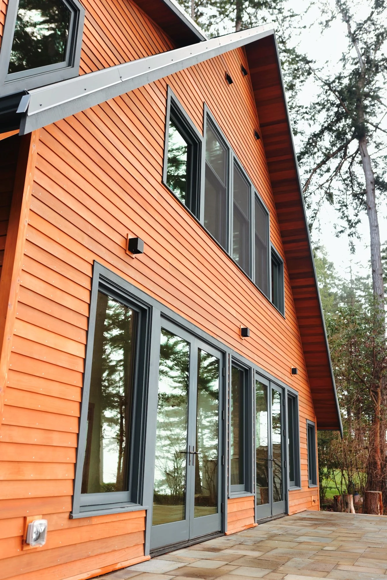 A modern house with orange wooden siding, large gray-framed windows, and a small patio with stone tiles. Tall trees are visible in the background.