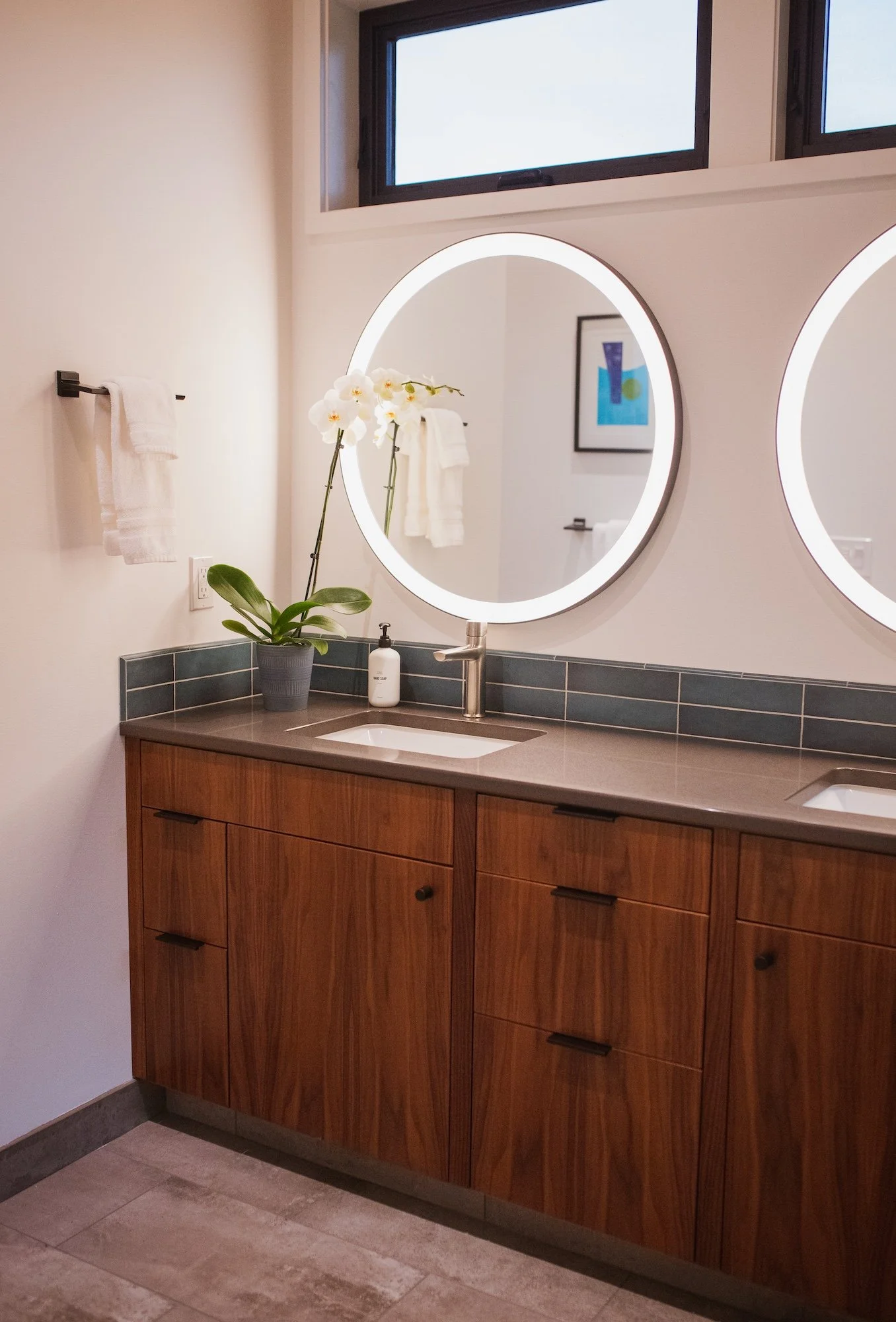 Bathroom vanity with a wooden cabinet, a white sink, a silver faucet, a potted orchid plant, a soap dispenser, and illuminated oval mirrors. There is a window above the mirrors, a towel rack with white towels on the wall, and a framed abstract art pi