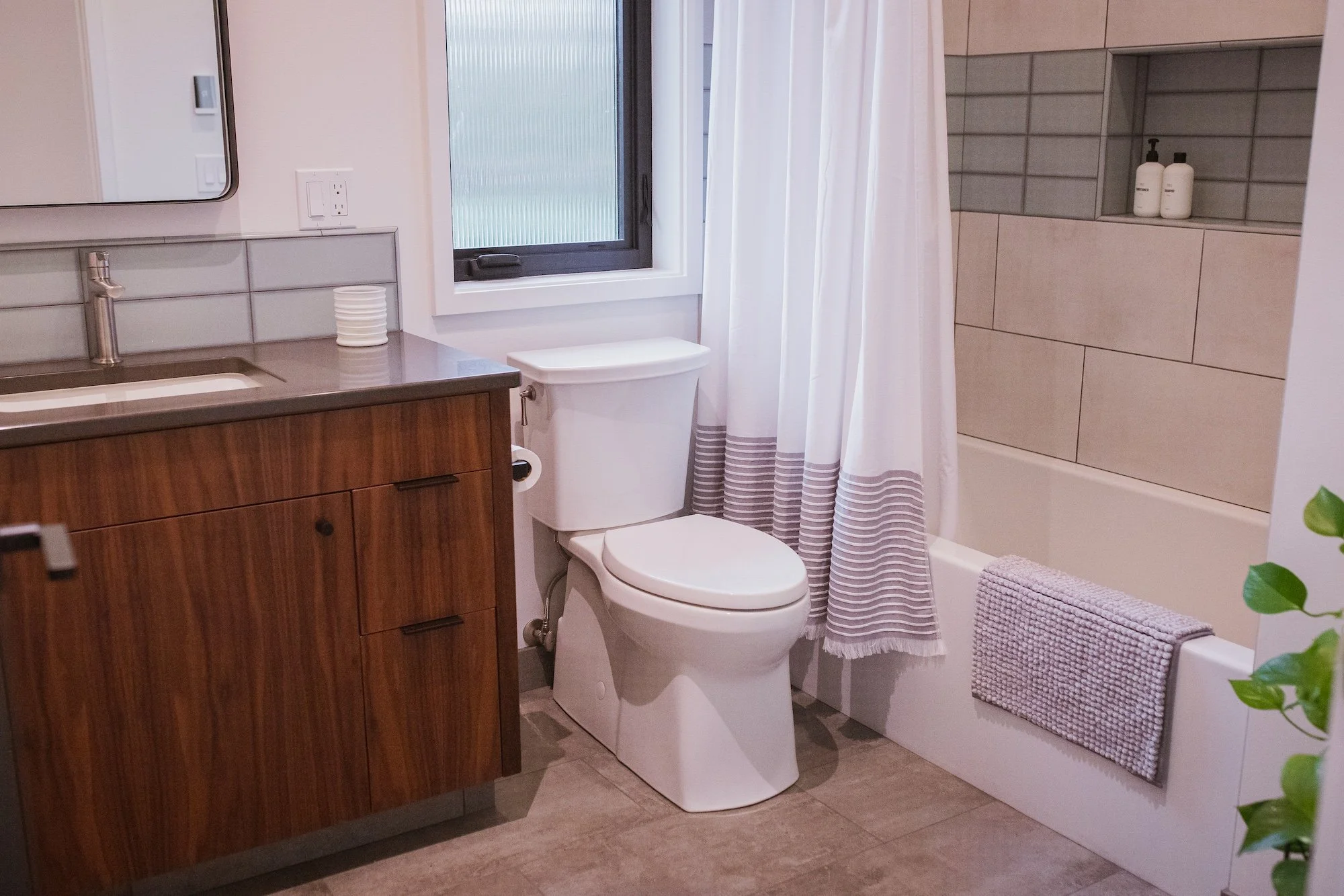 A modern bathroom with a wooden vanity, a white toilet, a window with white curtains, a gray bathtub with a tiled wall, and toiletries on a built-in shelf.