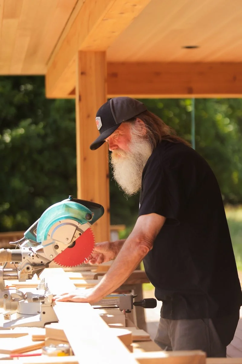 An elderly man with a long white beard, wearing a black baseball cap and black t-shirt, using a circular saw to cut wood outdoors.