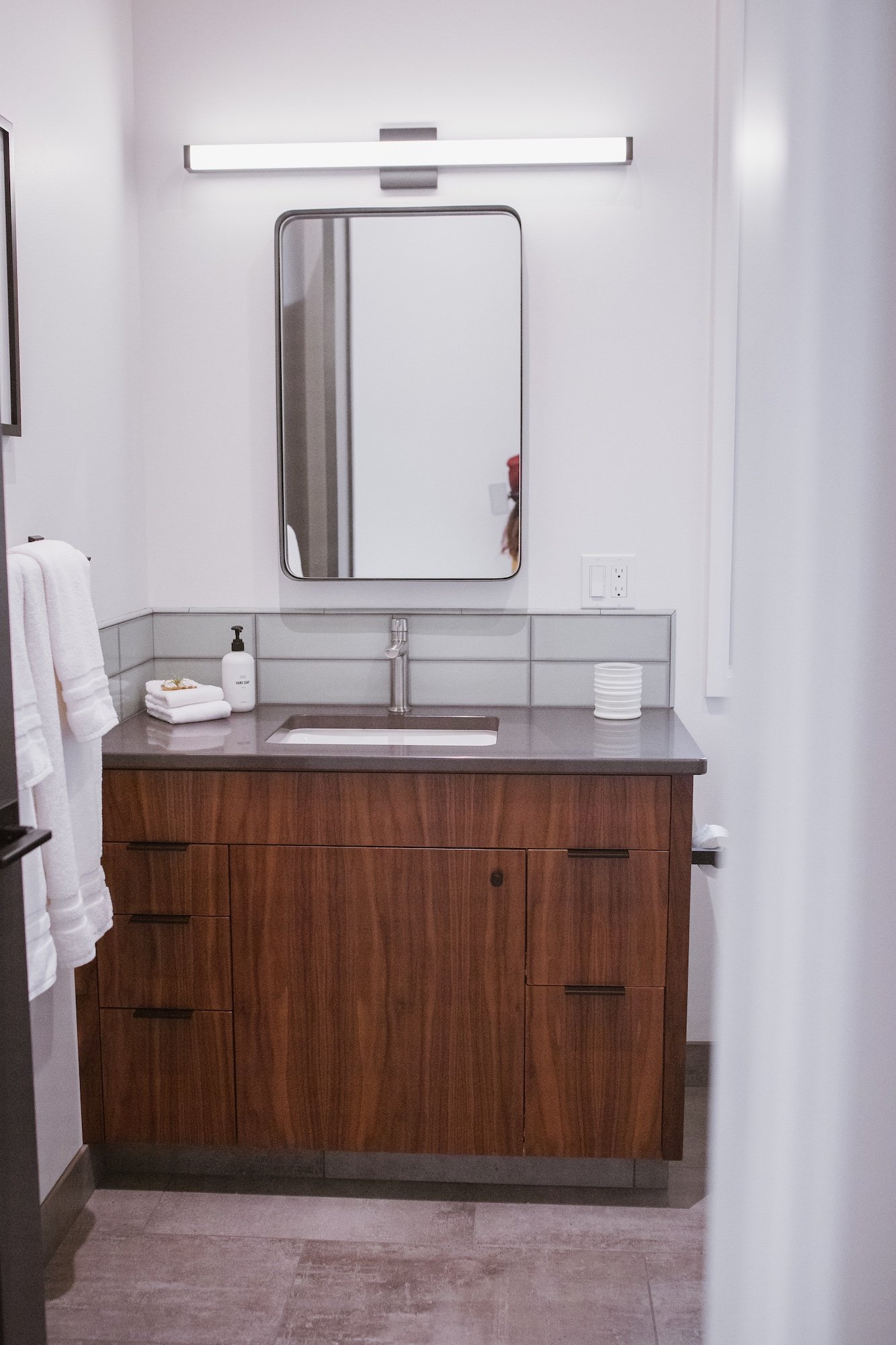 A modern bathroom vanity with a wooden cabinet, a gray countertop, a rectangular under-mount sink, and a wall-mounted mirror above. There is a light fixture above the mirror, and a soap dispenser, folded towels, and a small container are on the count