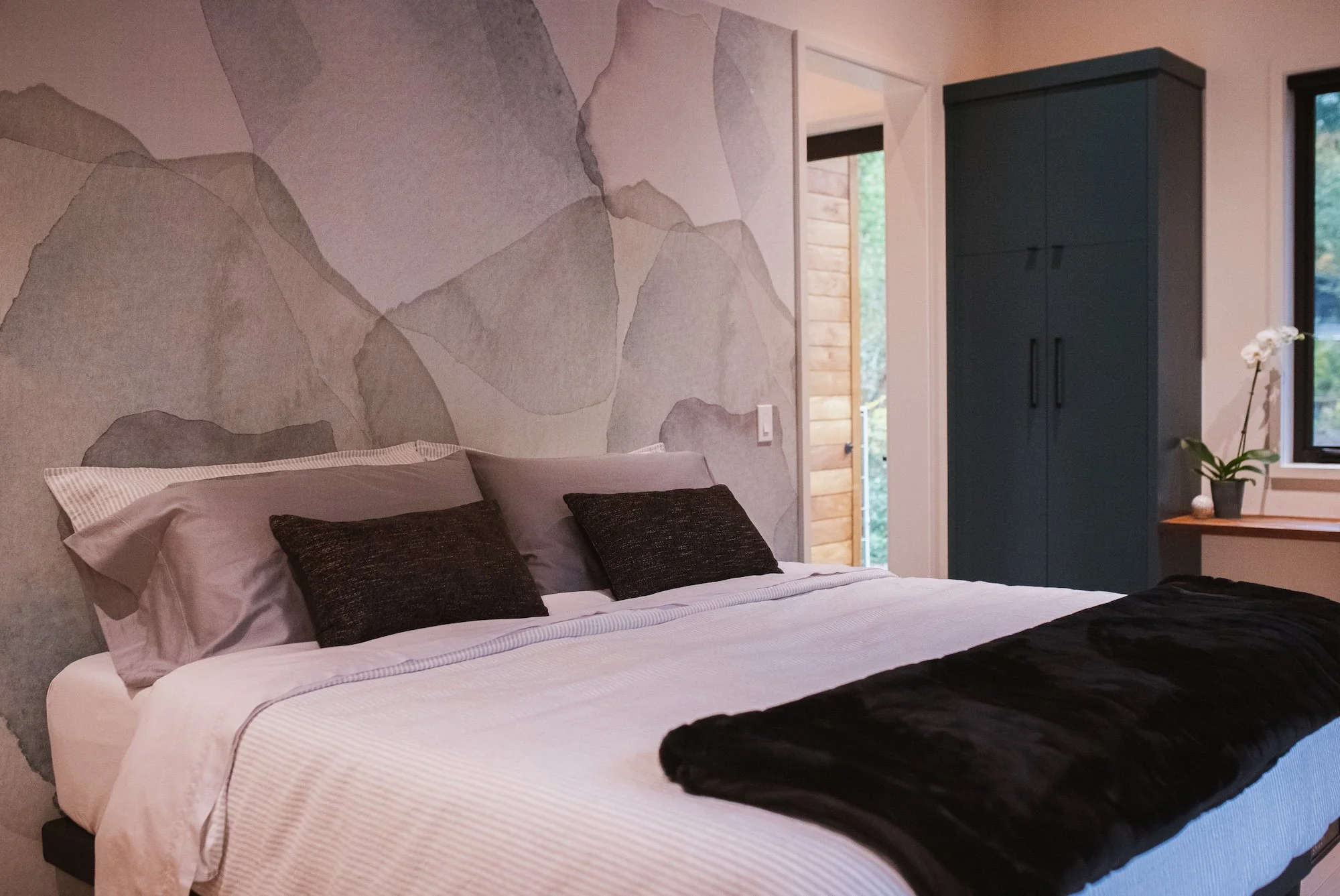 A bedroom with a bed, dark pillows, and a black throw blanket. The bed is against a wall with a mountain landscape mural. Next to the bed is a black wardrobe, a window, and a small wooden table with a potted orchid.