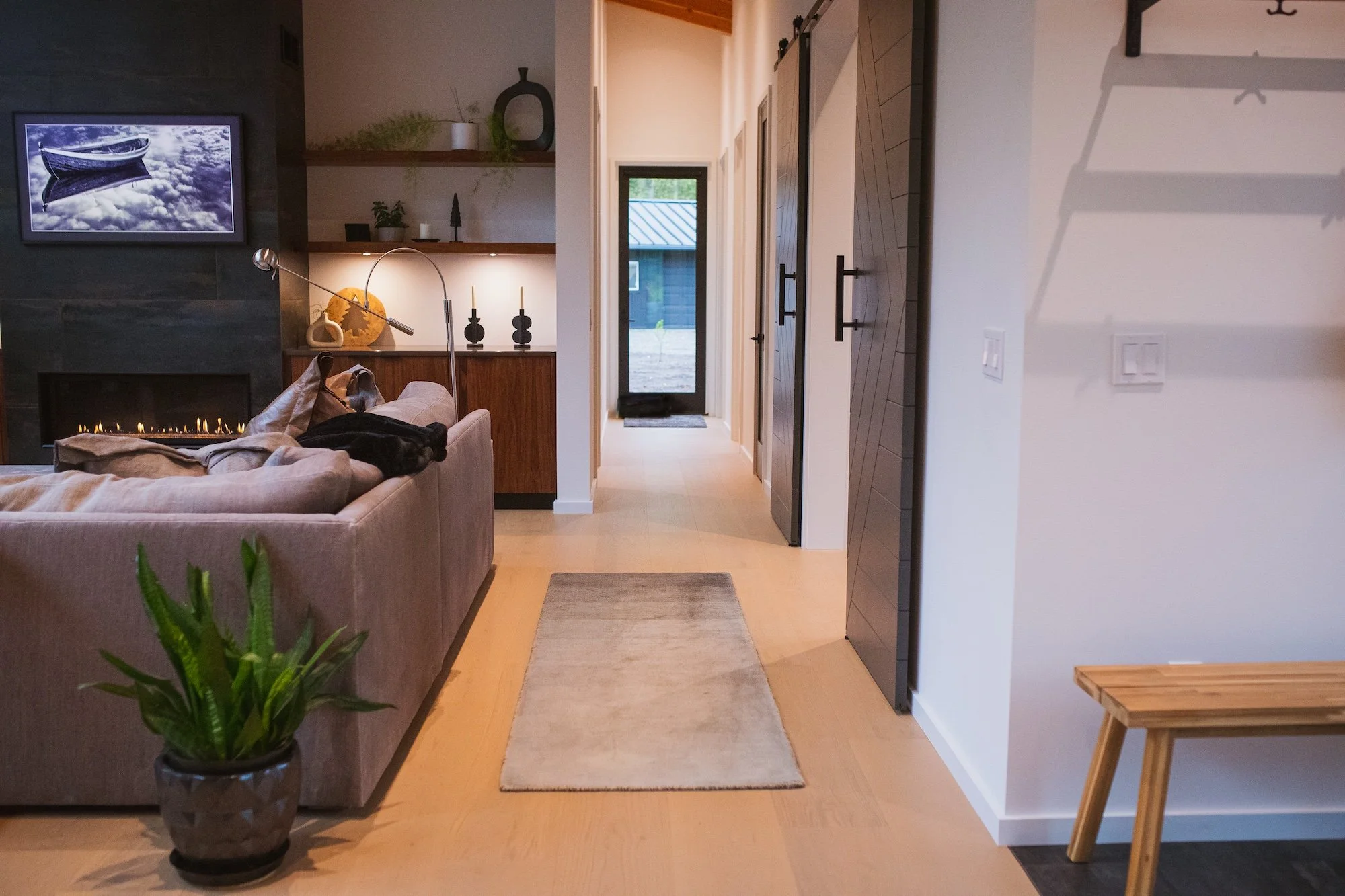 Interior view of a modern living space with a beige sofa, a potted plant, open shelves with decorative items, a fireplace, and a hallway with closed barn doors leading to other rooms.