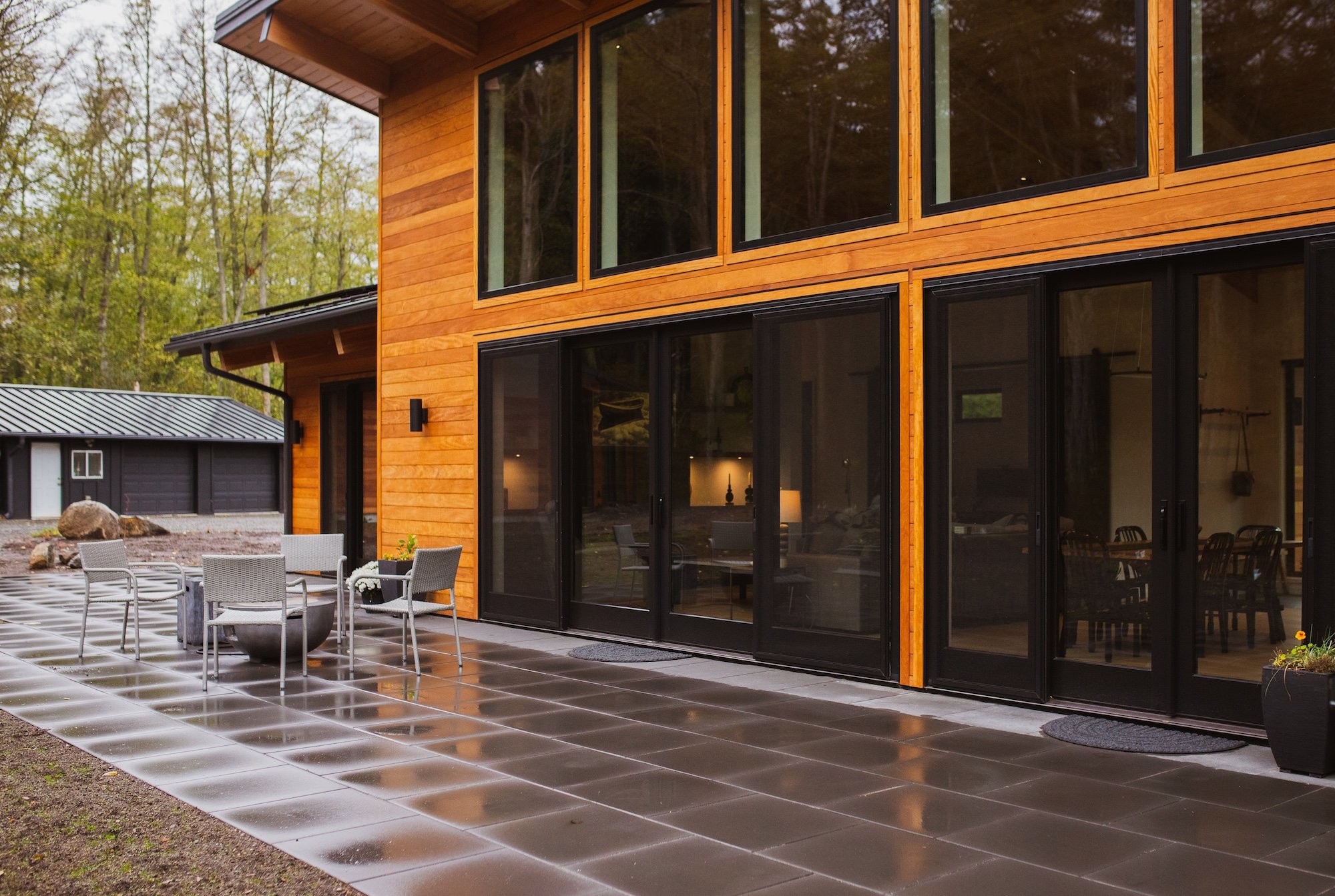 Modern two-story house with large black-framed glass doors and windows, wooden exterior, patio furniture, and a wet tiled patio area, surrounded by trees.
