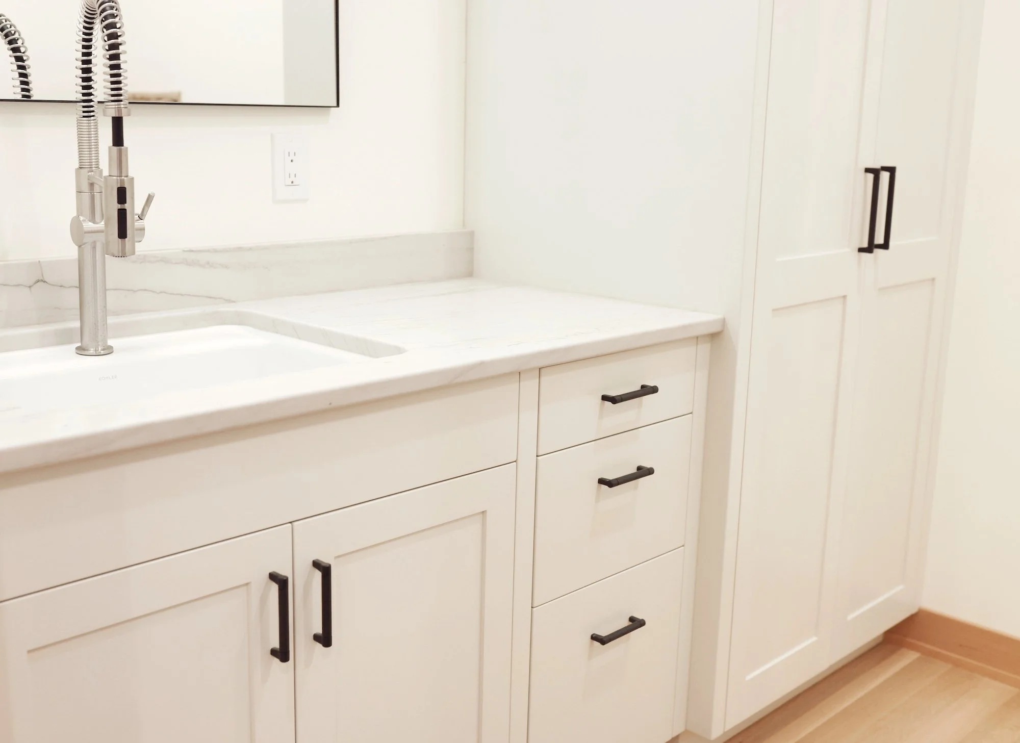 White kitchen cabinet with black handles, a marble countertop, and a stainless steel faucet for the sink, with a mirror and electrical outlet on the wall.