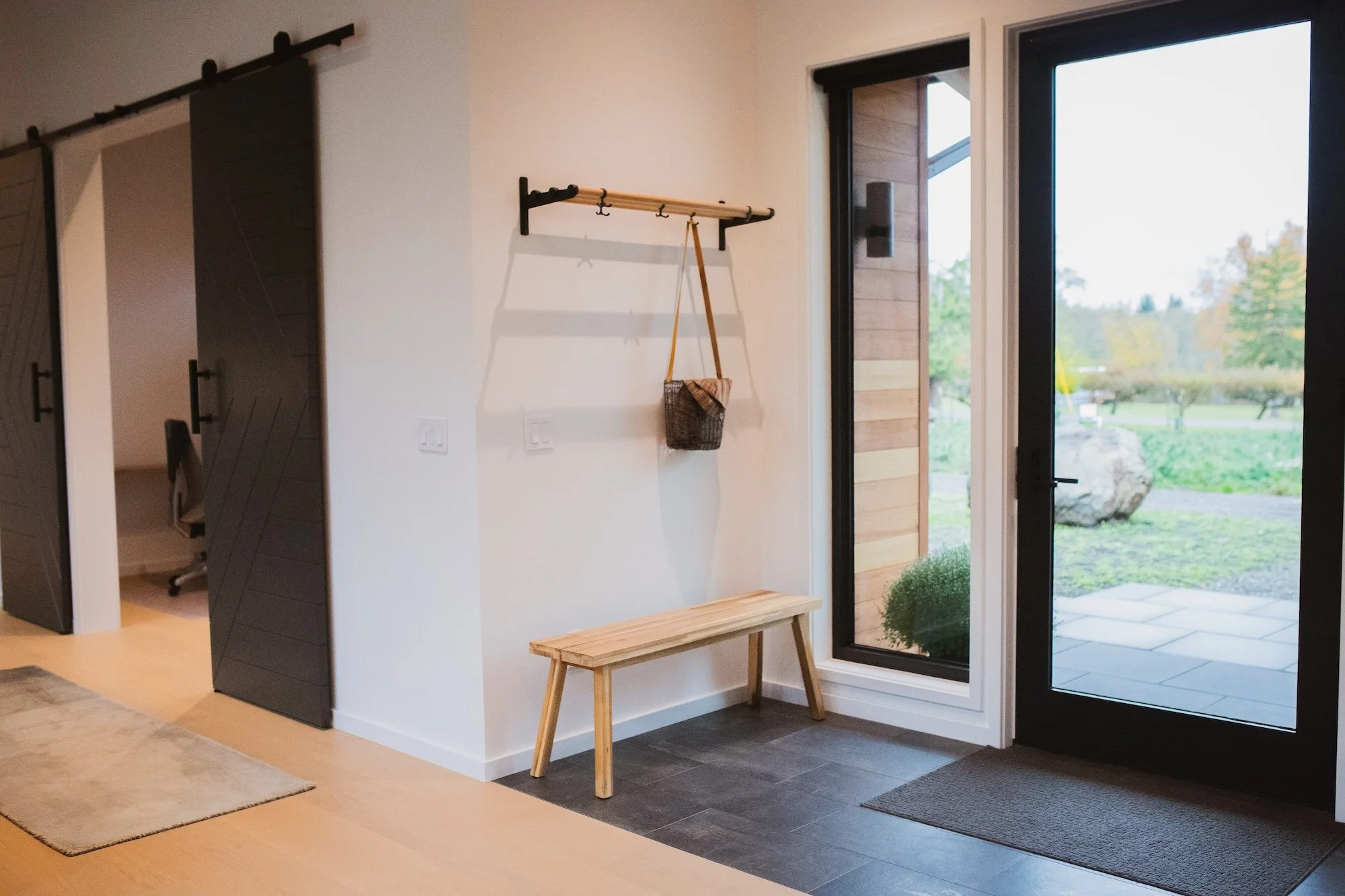 Interior entryway with a windowed door, a wooden bench, a wall-mounted coat rack with a basket hanging from one hook, and a bench beneath it. Outside the window, trees and a large rock are visible.