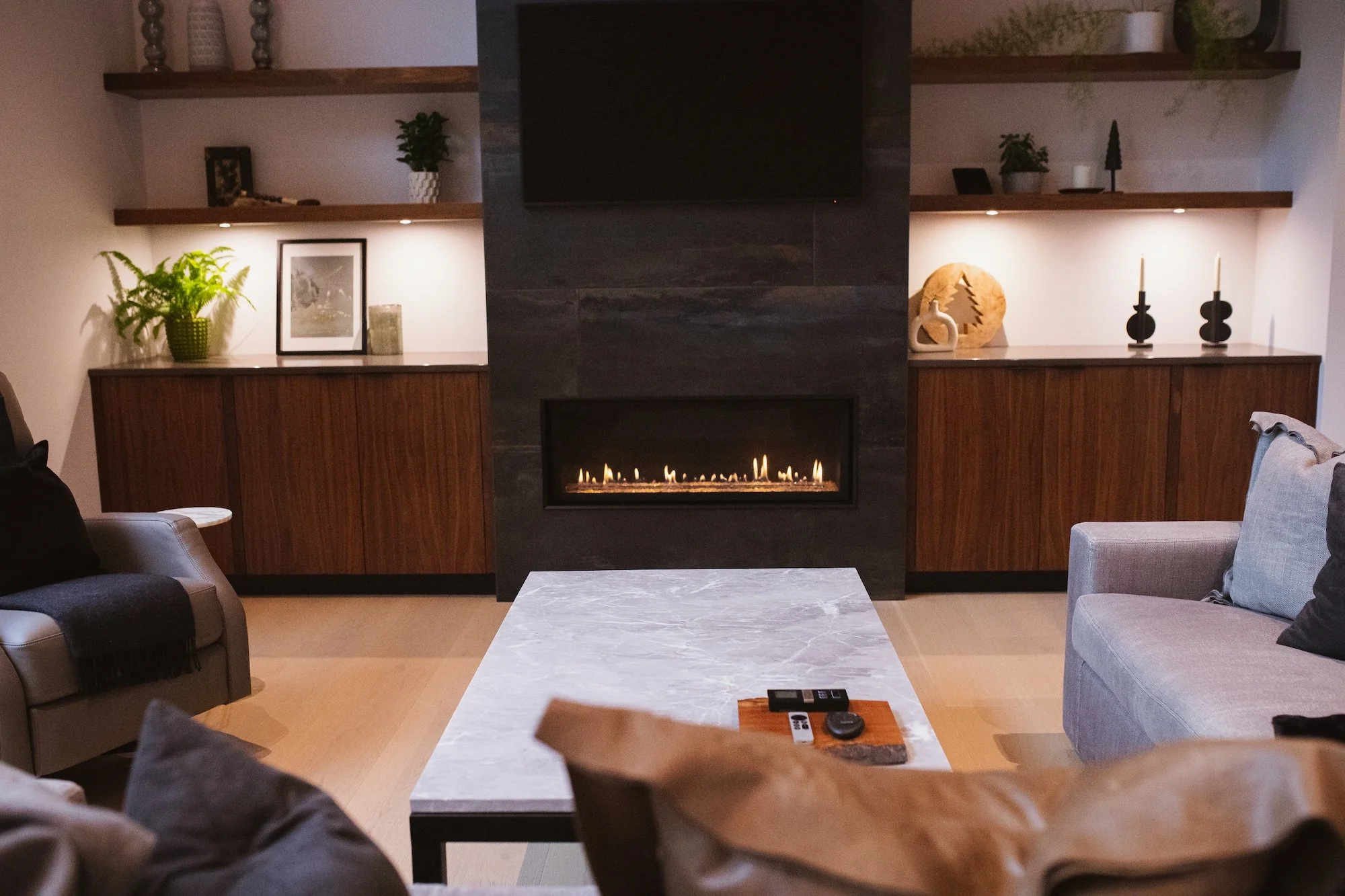 Living room with a modern black fireplace, refrigerated with a flame, between two wooden sideboards with decorative objects and lamps. Shelves with plants and decor items above the sideboards. Gray and beige sofas and chairs, a marble coffee table wi