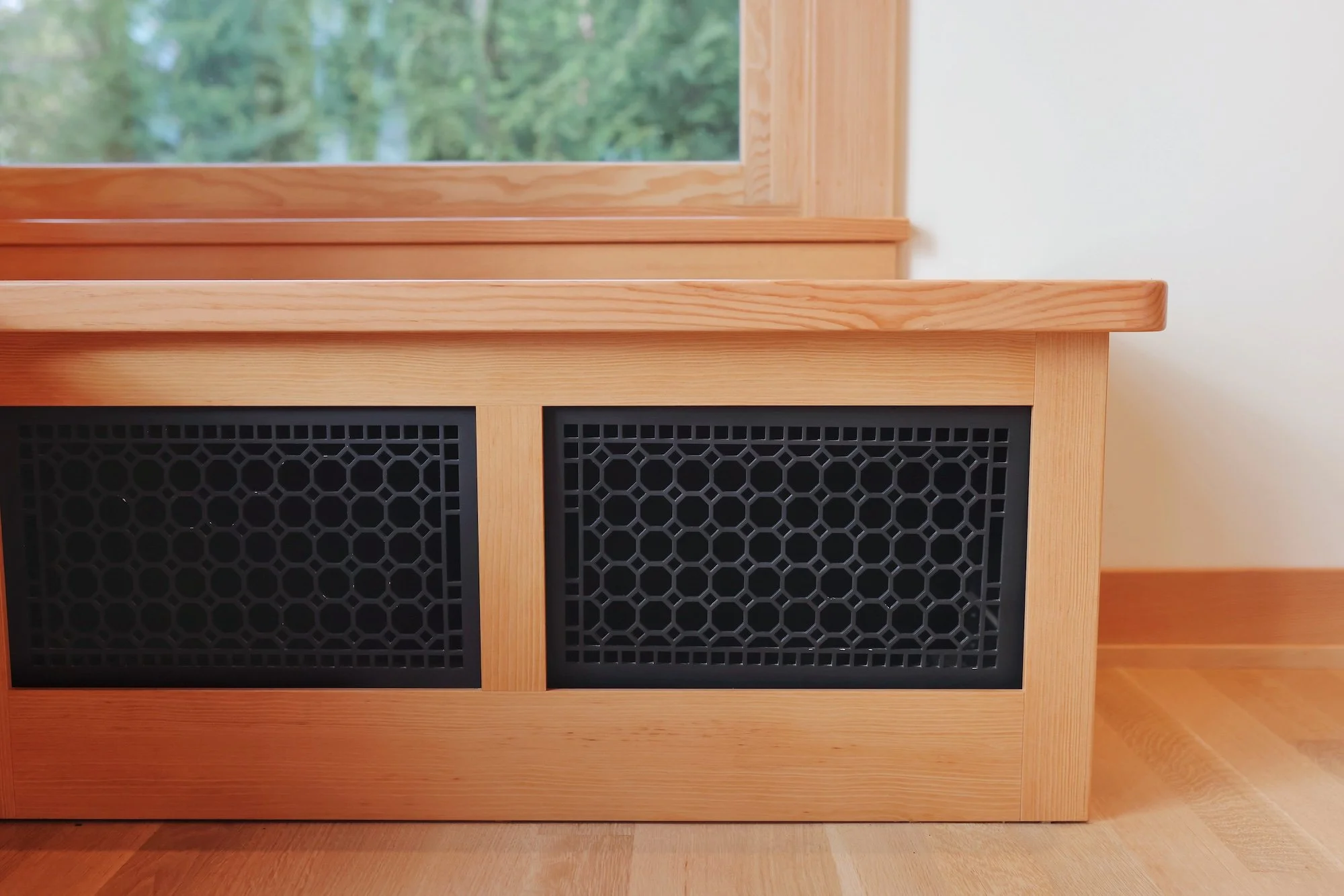 A wooden cabinet with black vented panels on the doors, positioned in front of a wall with a window showing trees outside.