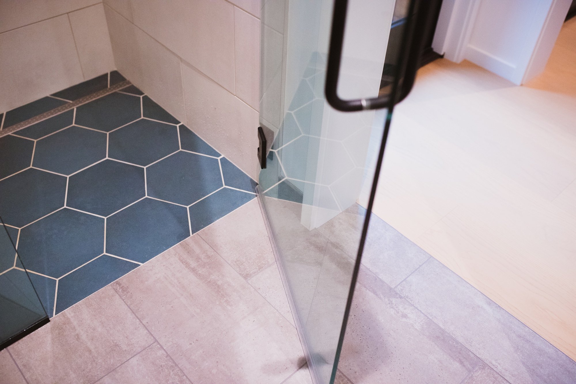 Interior view of a bathroom or changing room with a partially open glass door, hexagonal blue tiles on the floor, and off-white tiles on the wall. There is a black handle on the glass door and a black hook on the wall.