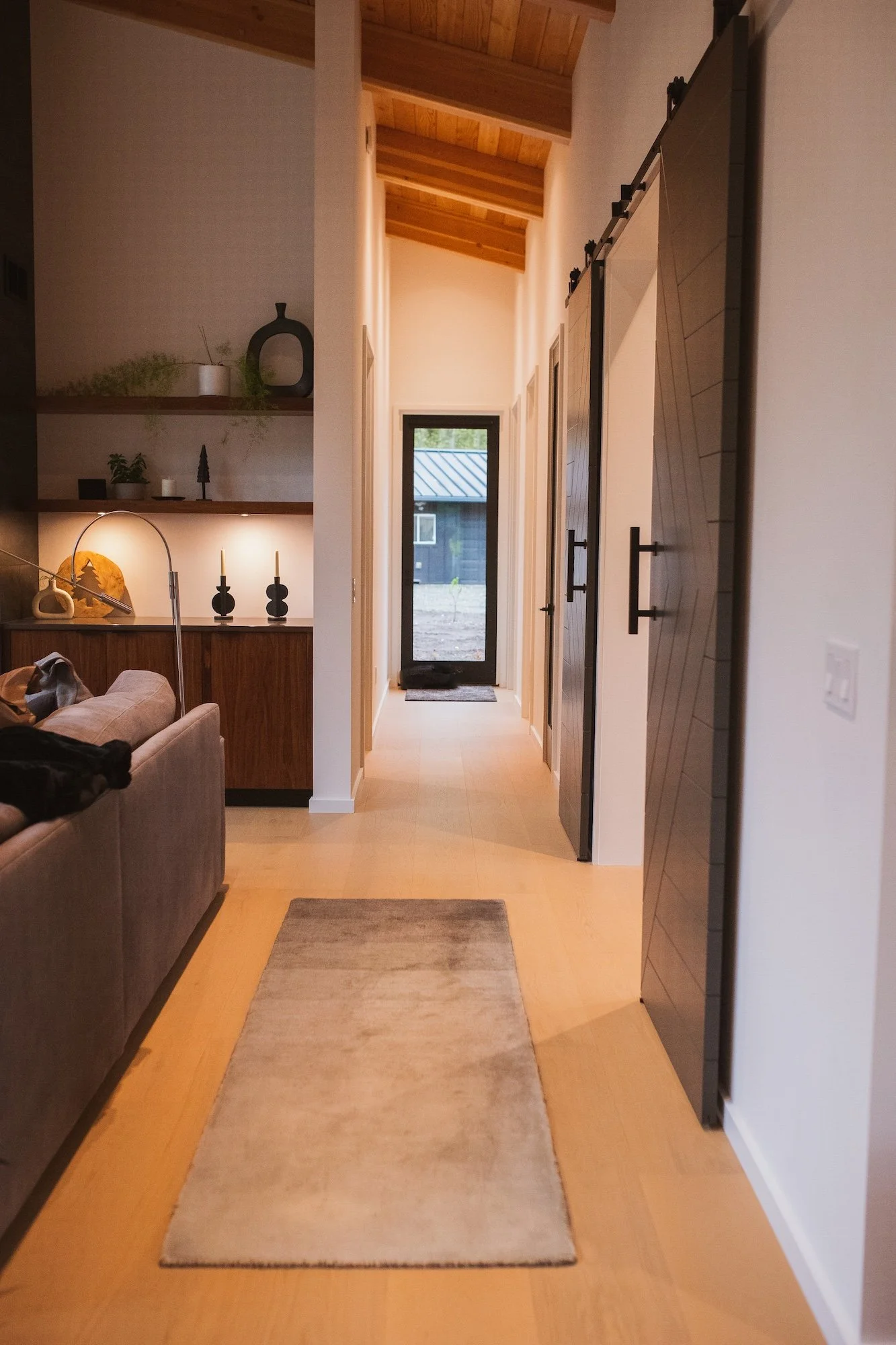 A hallway with a light-colored wooden floor, a beige runner rug, and white walls. There is a sofa on the left side, a small coffee table, and decorative items on the shelves. At the end of the hallway, there is a black door with a window and greenery