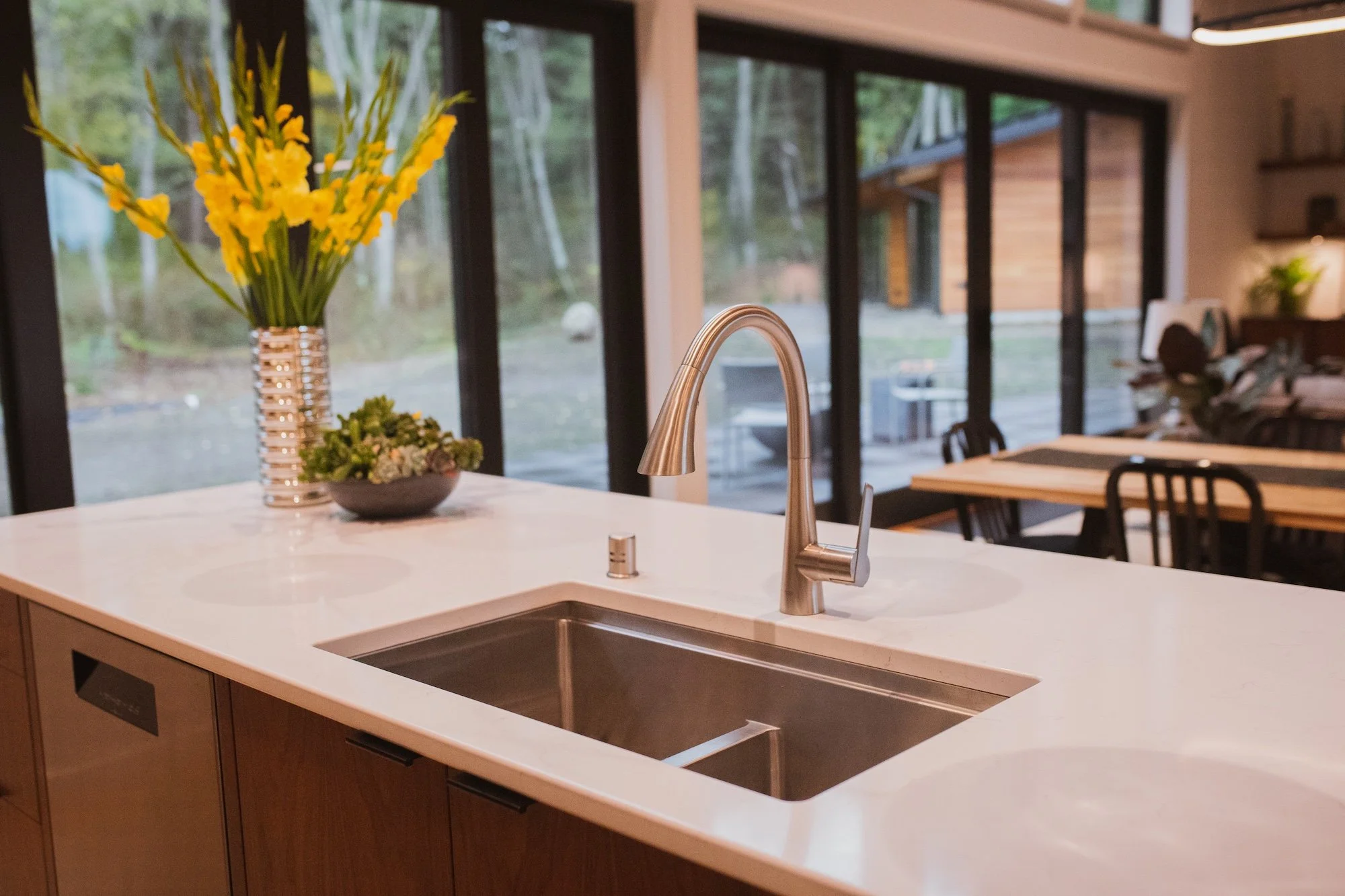 A modern house kitchen with a white countertop, a stainless steel sink with a curved faucet, a bowl of small succulents, and a tall glass vase filled with bright yellow flowers. Large windows show a wooded outdoor landscape.