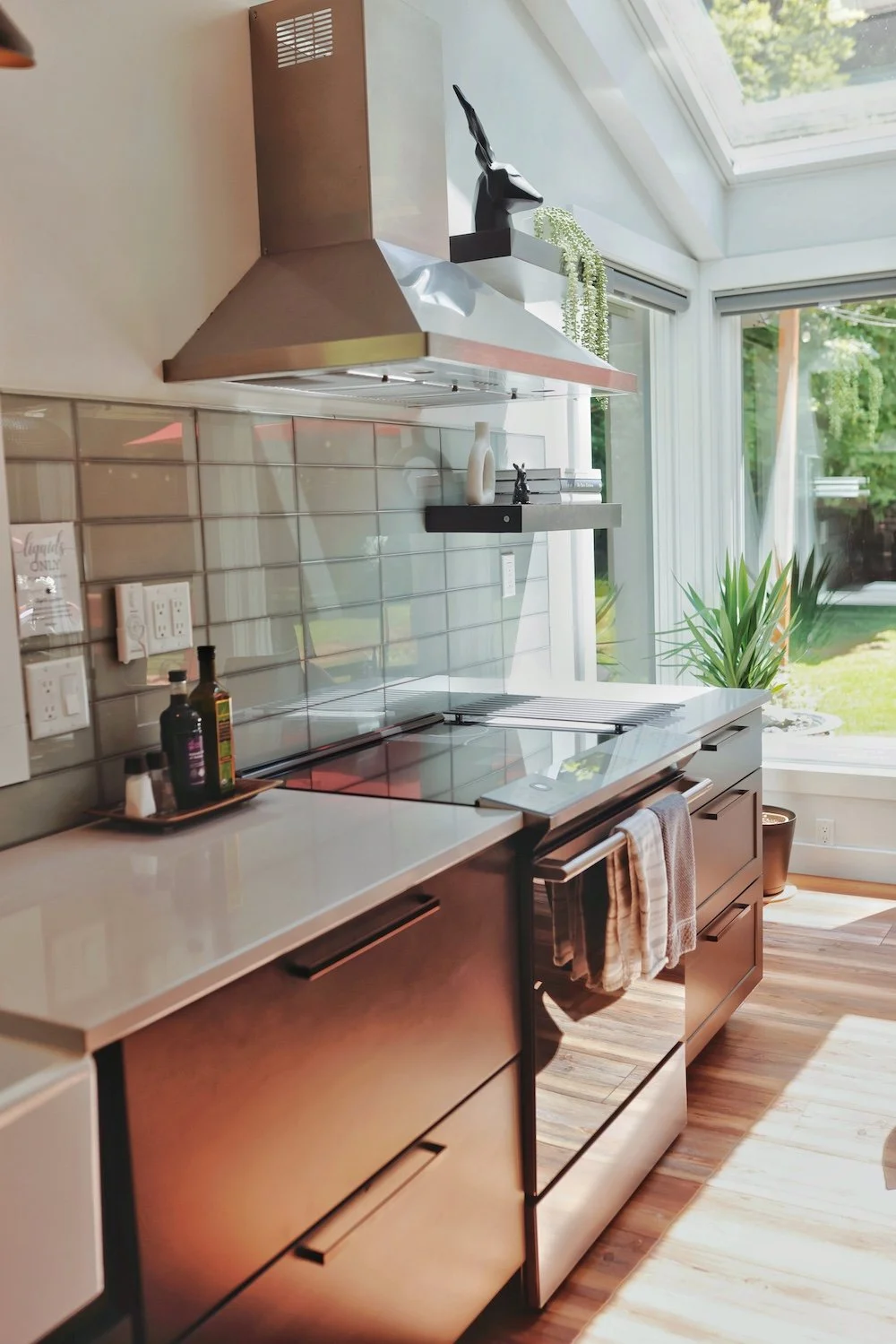 Modern kitchen with stainless steel appliances and large windows, featuring a countertop with condiments, a stove, and decorative decor on shelves, with sunlight and greenery outside.