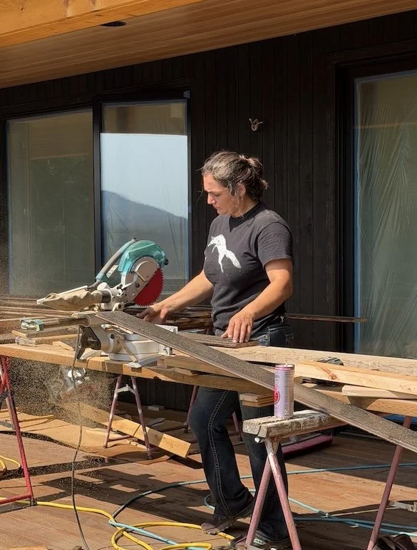 A woman cutting wood with a miter saw on a workbench outside a house with large windows.