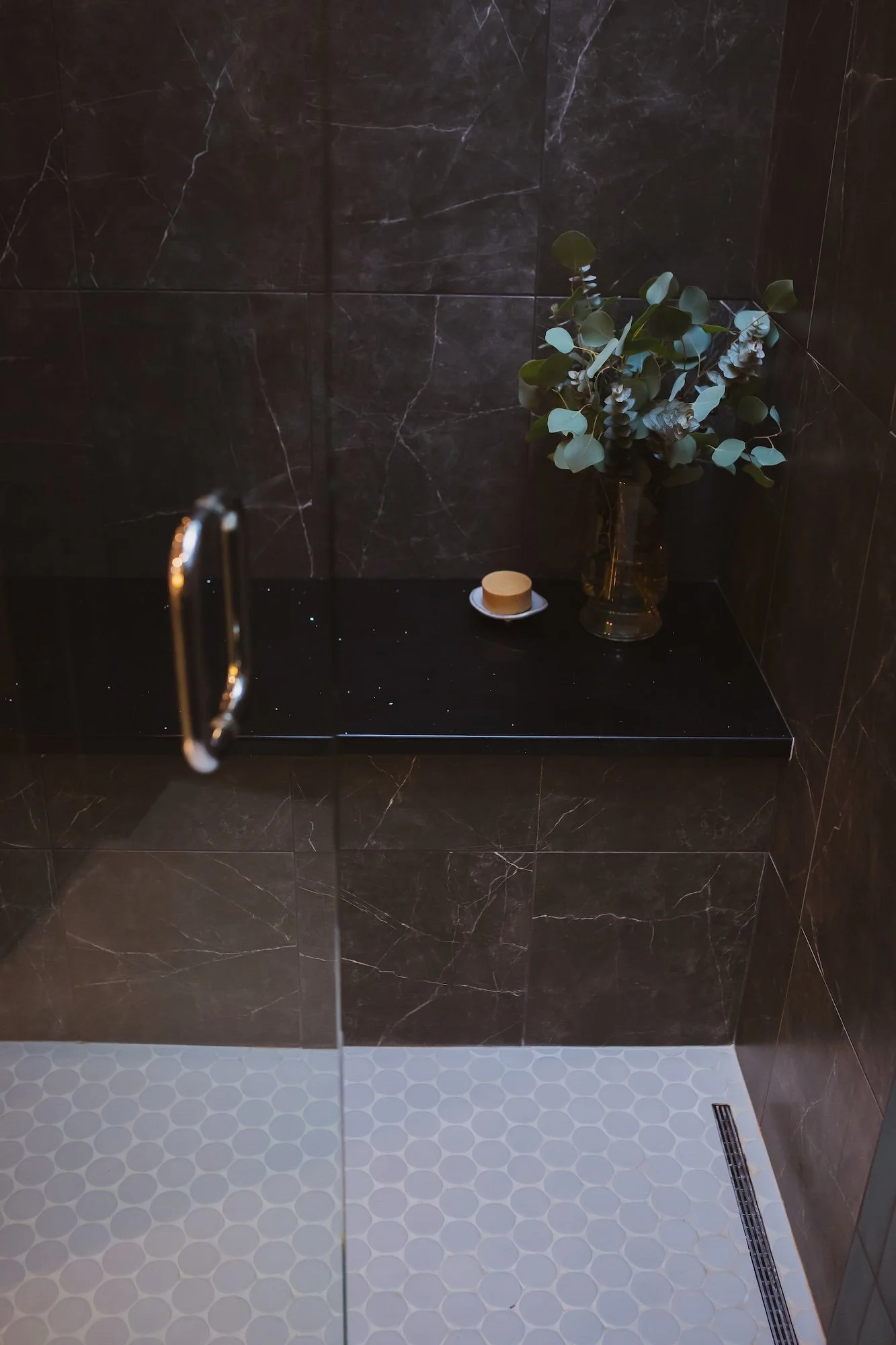 View of a shower with dark marble tiled walls, a vase with green eucalyptus leaves and a beige soap on a small white dish, and a floor with white hexagonal tiles and a linear drain.