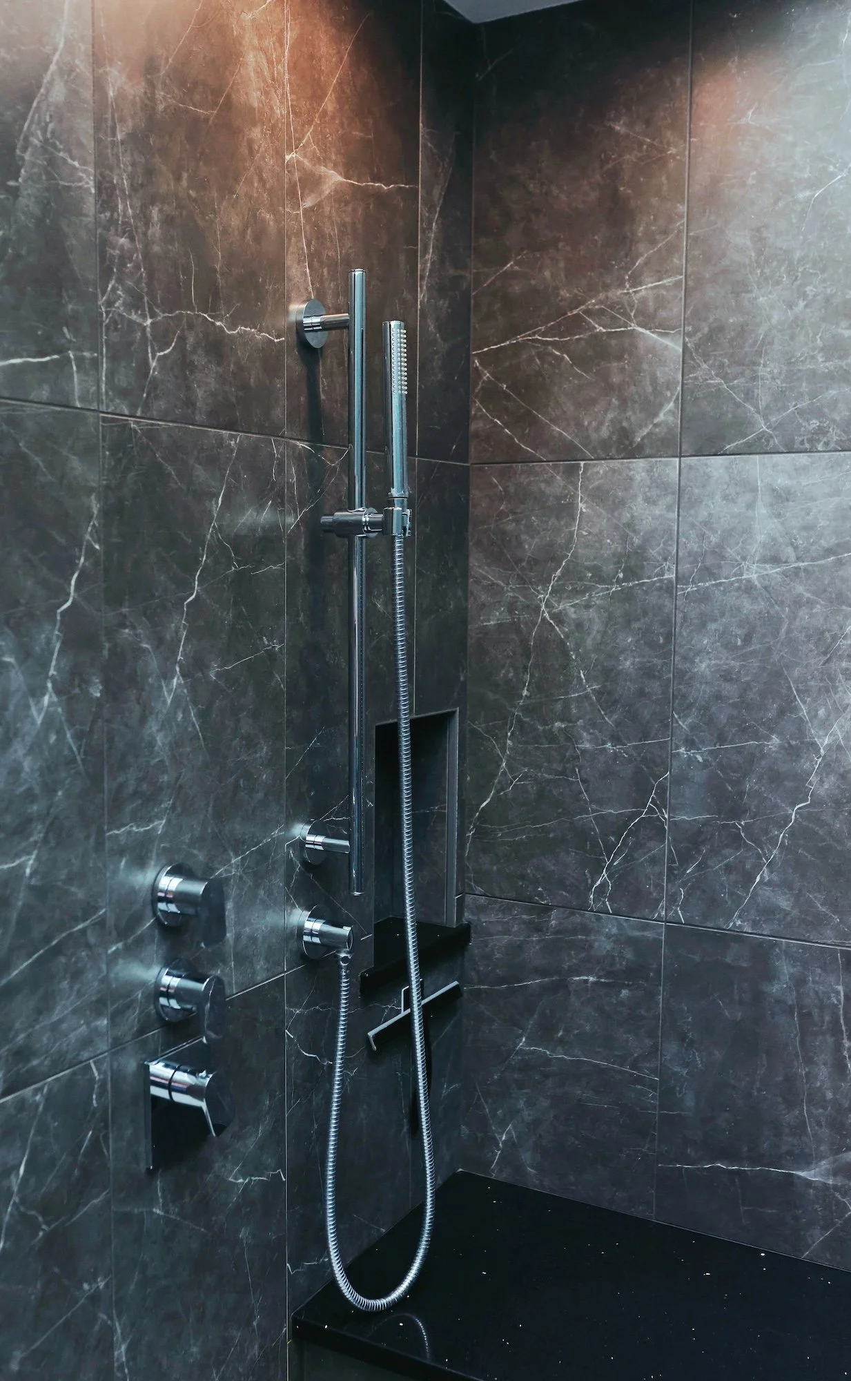 Modern shower area with black marble walls, multiple shower controls, a handheld shower with a flexible hose, a fixed shower bar, and a black bench at the corner.