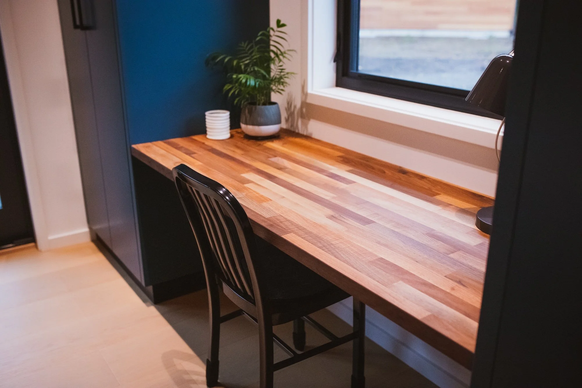 A wooden kitchen counter next to a window with a view outside. There are some plants and a stack of white plates on the counter, with a black chair partially visible in front.