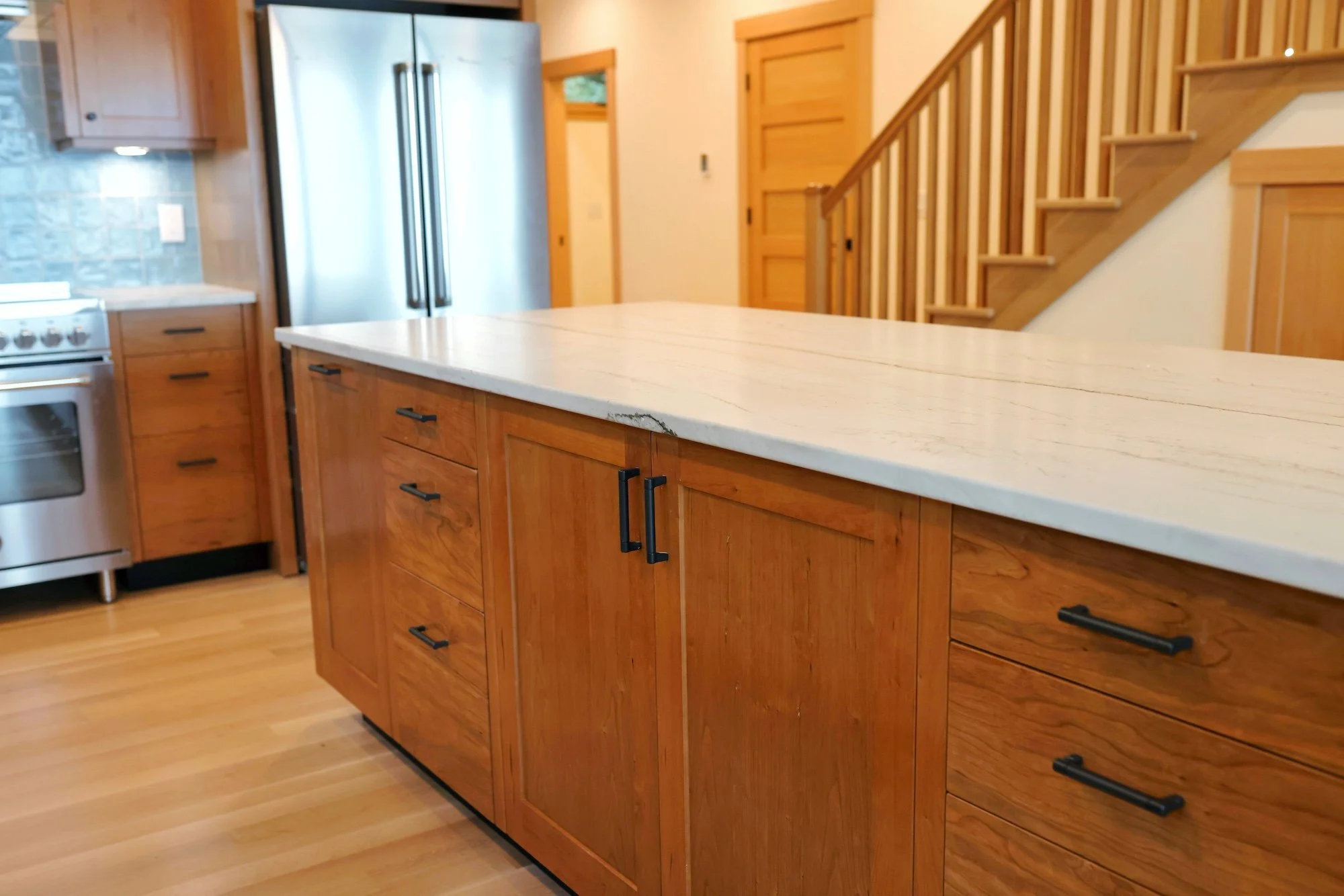 A kitchen with wooden cabinets and a white marble countertop, stainless steel refrigerator, wooden staircase, and hardwood floors.