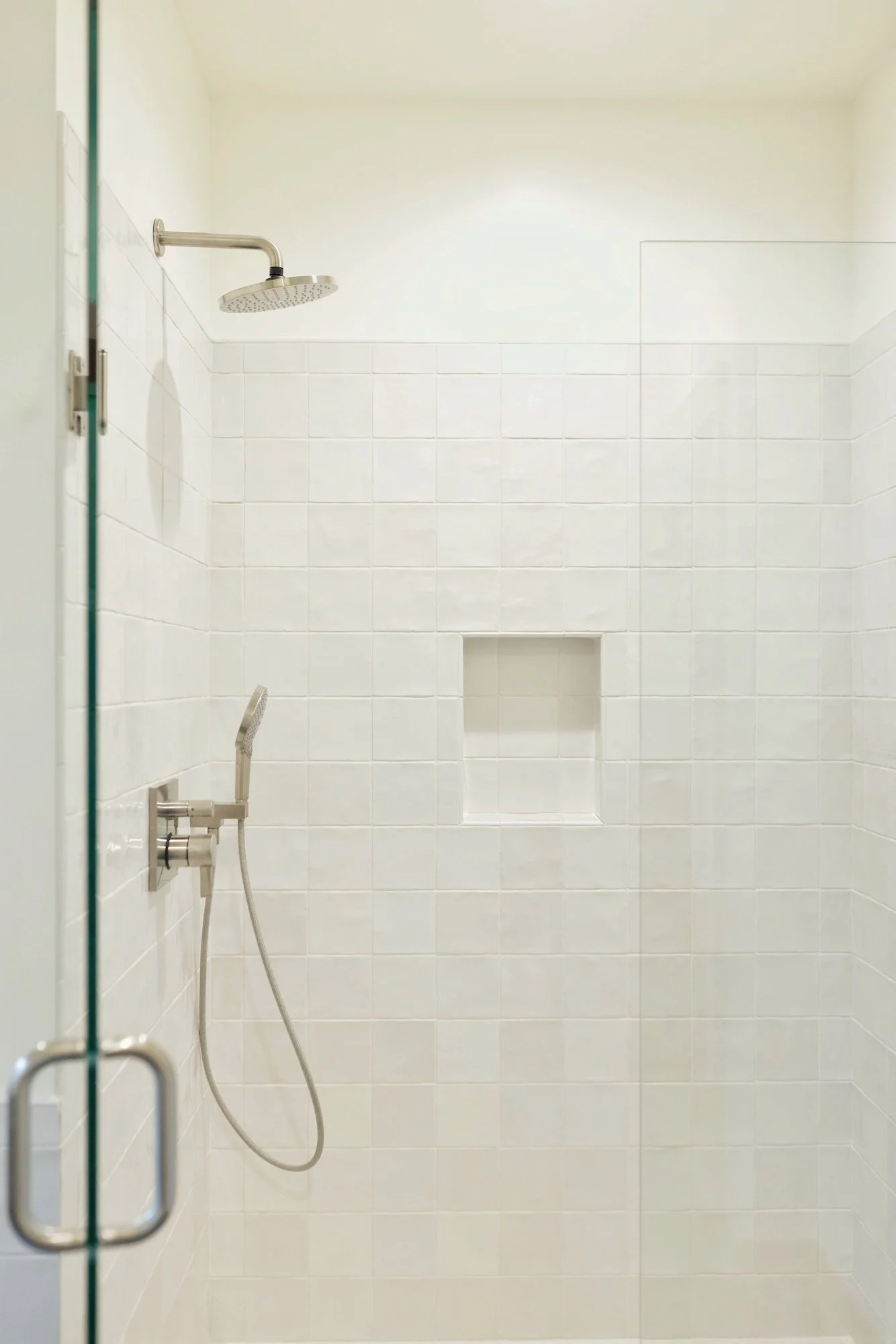 Shower area with a large rainfall showerhead, handheld shower, white tiled walls, built-in niche, and glass door.
