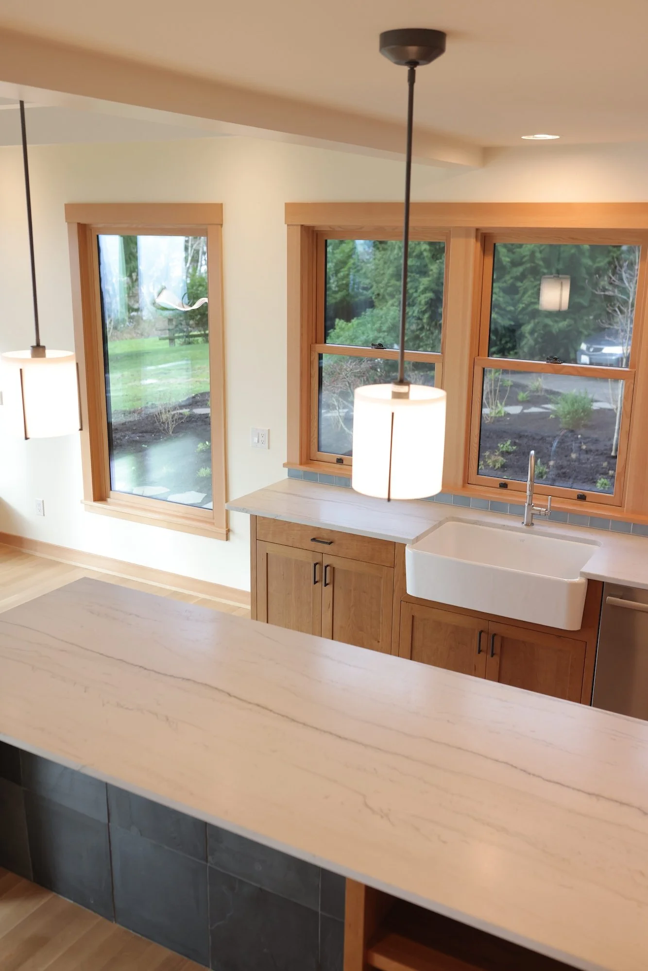 A kitchen with light wood cabinets, a white farmhouse sink, large windows showing a backyard garden, pendant lights, and a marble countertop.