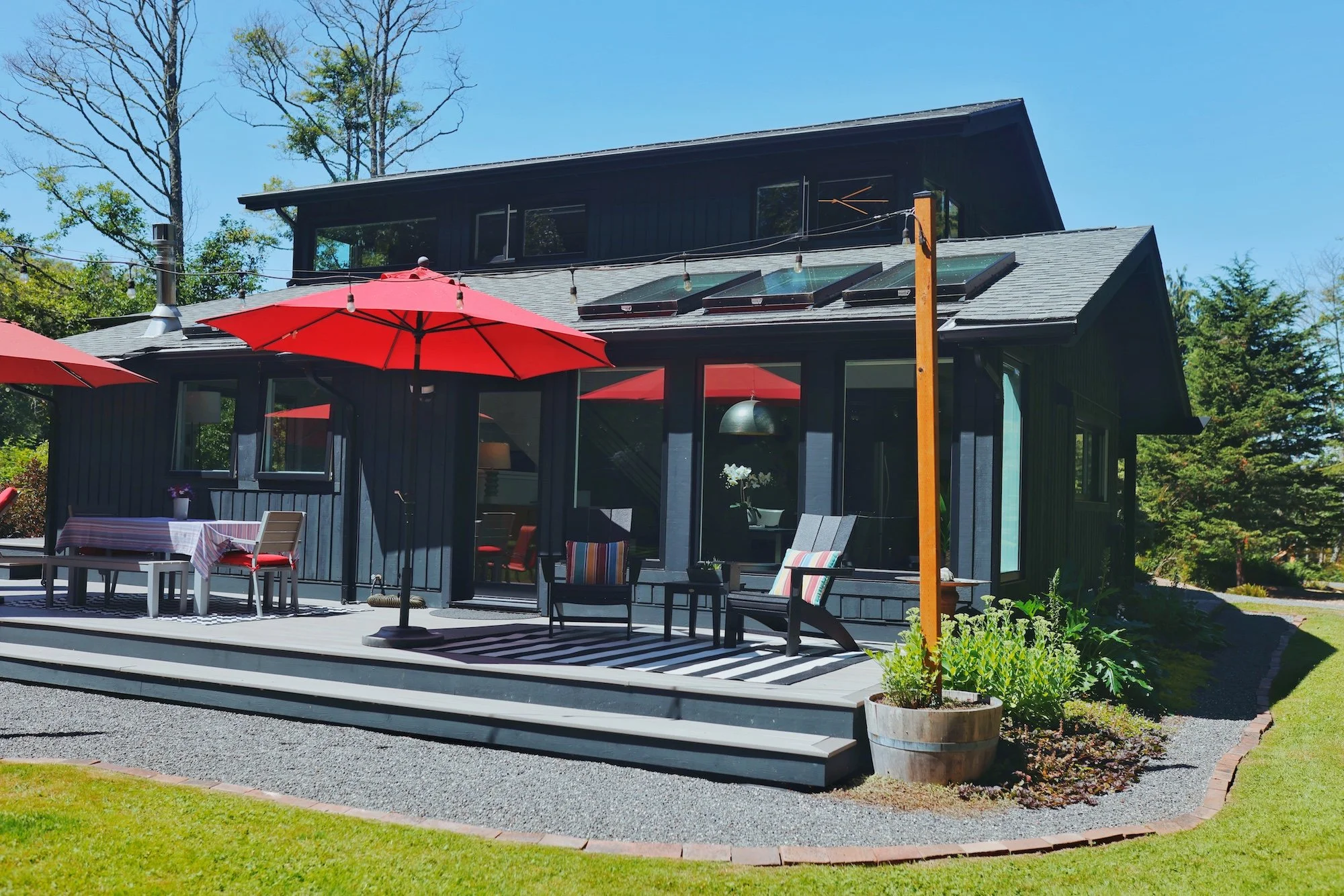 A modern black house with a deck and multiple large windows, surrounded by trees and a well-maintained lawn. Red patio umbrellas and outdoor furniture are on the deck, including a table with a checkered tablecloth, chairs, and potted plants.