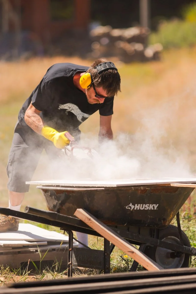 A man wearing yellow ear protection and yellow gloves using a chainsaw to cut a piece of wood on a Husky wheelbarrow outdoors on a sunny day.