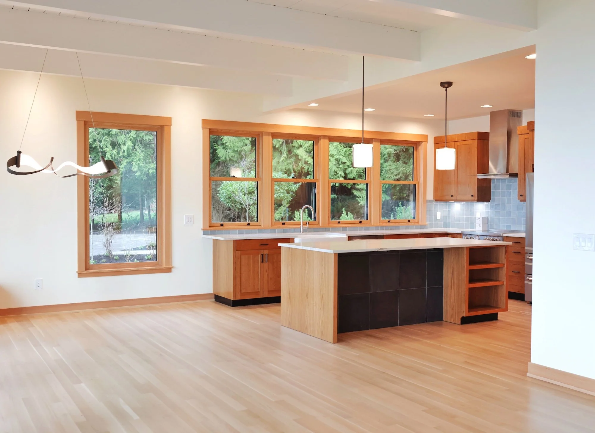 Modern kitchen with wooden cabinets, a kitchen island, and large windows showing greenery outside.