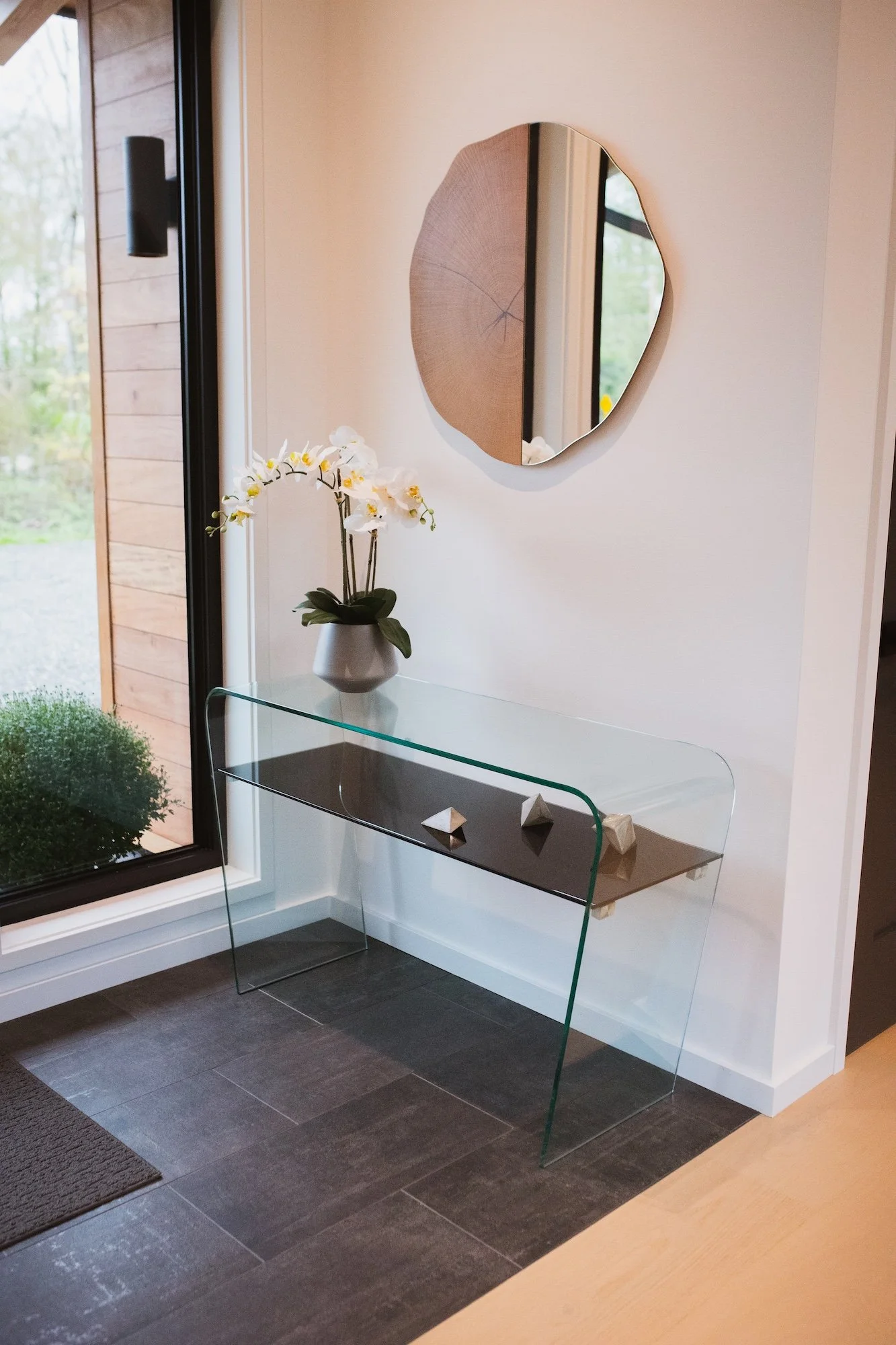 A modern living space with a glass console table holding a white orchid plant, a round mirror on the wall, and a view of outdoor greenery through a large window.