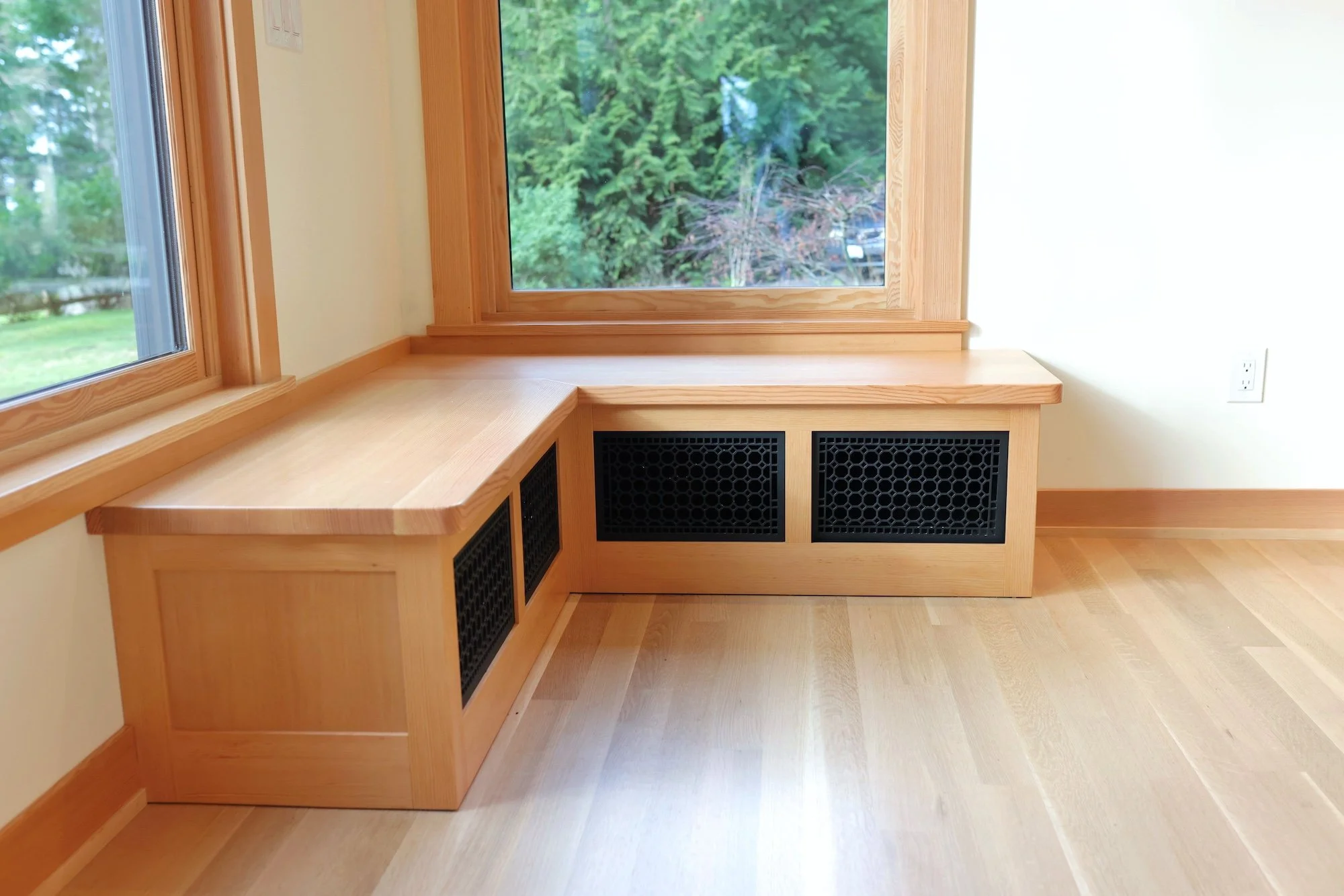 A corner of a room with wooden window frames, a window seat, and a heater cover.