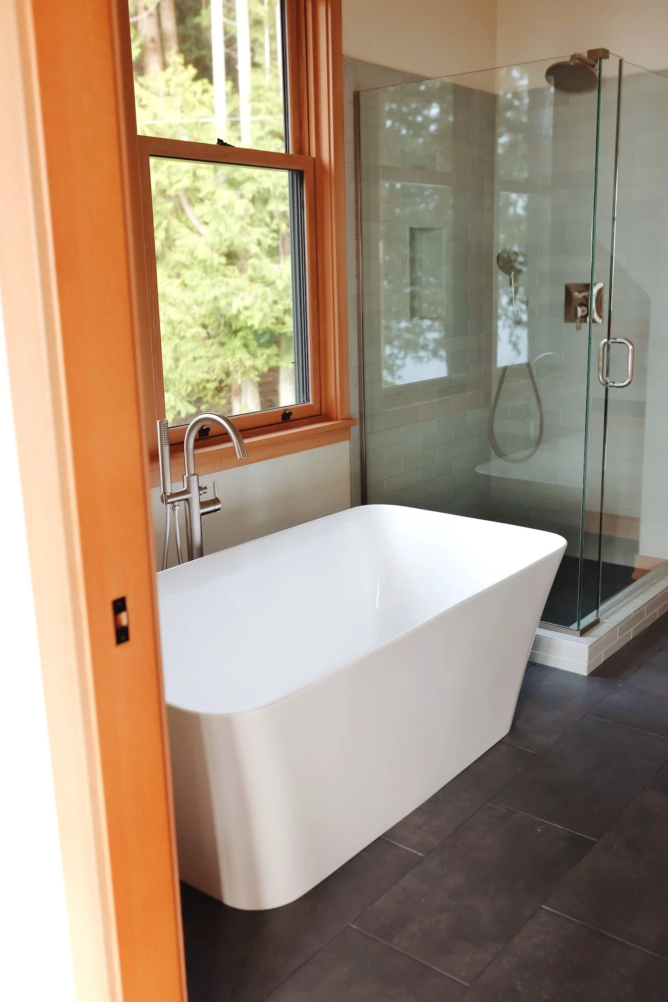 View of a modern bathroom with a white freestanding bathtub, a glass-enclosed shower with gray tiles, a wooden window with a view of greenery outside, and a dark gray floor.