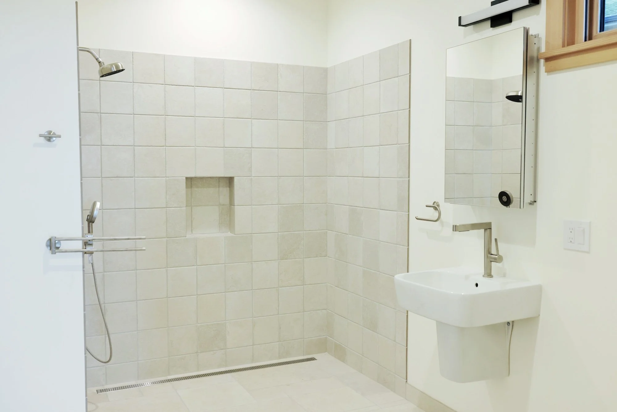 A clean, modern bathroom with a tiled shower area, a small sink, a mirror, and safety grab bars.